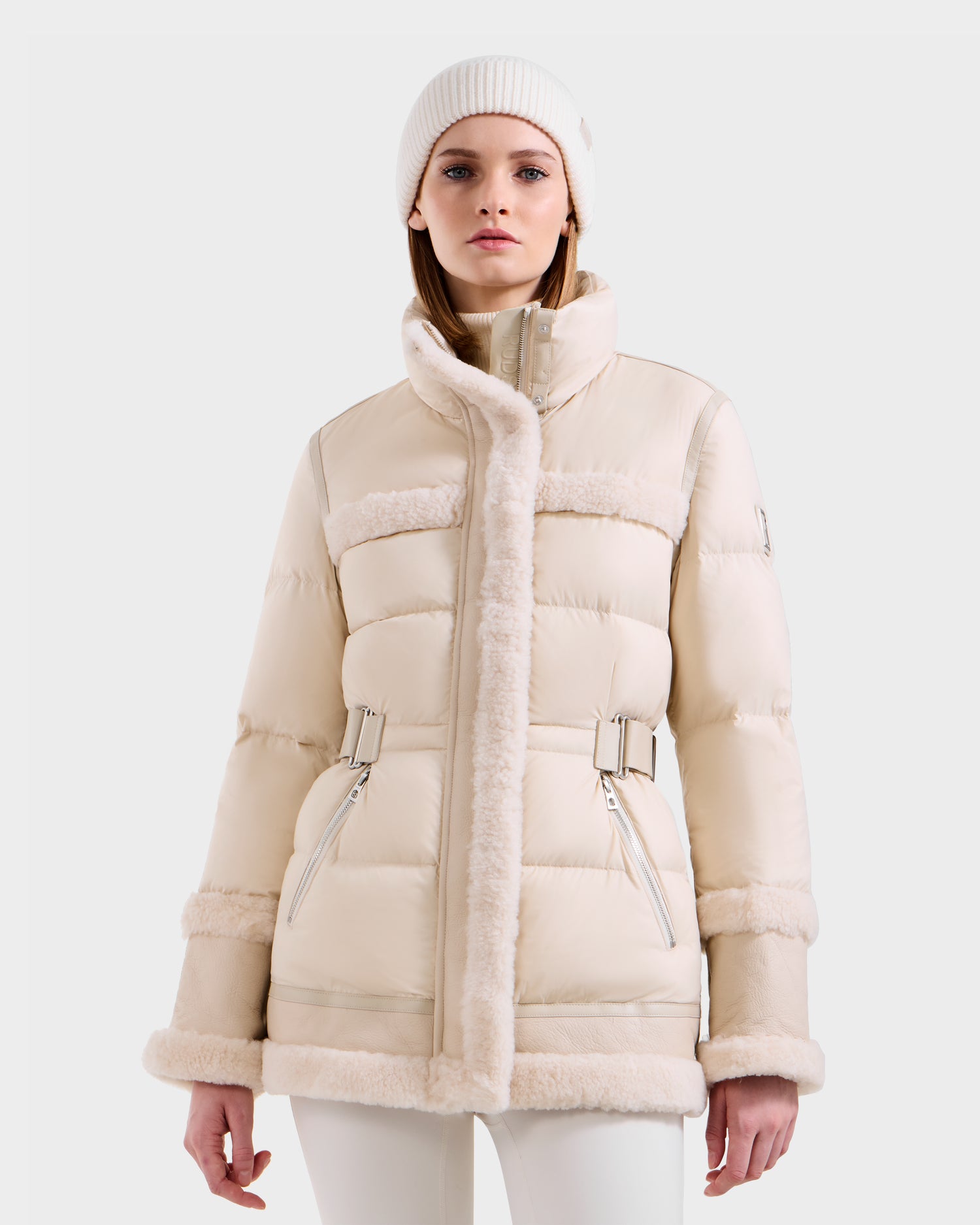 A woman wears the Rudsak TESSA - 8125615 Vanilla down puffer with shearling trim, zipped and belted, styled with a white knit beanie and white pants, standing against a plain light background.
