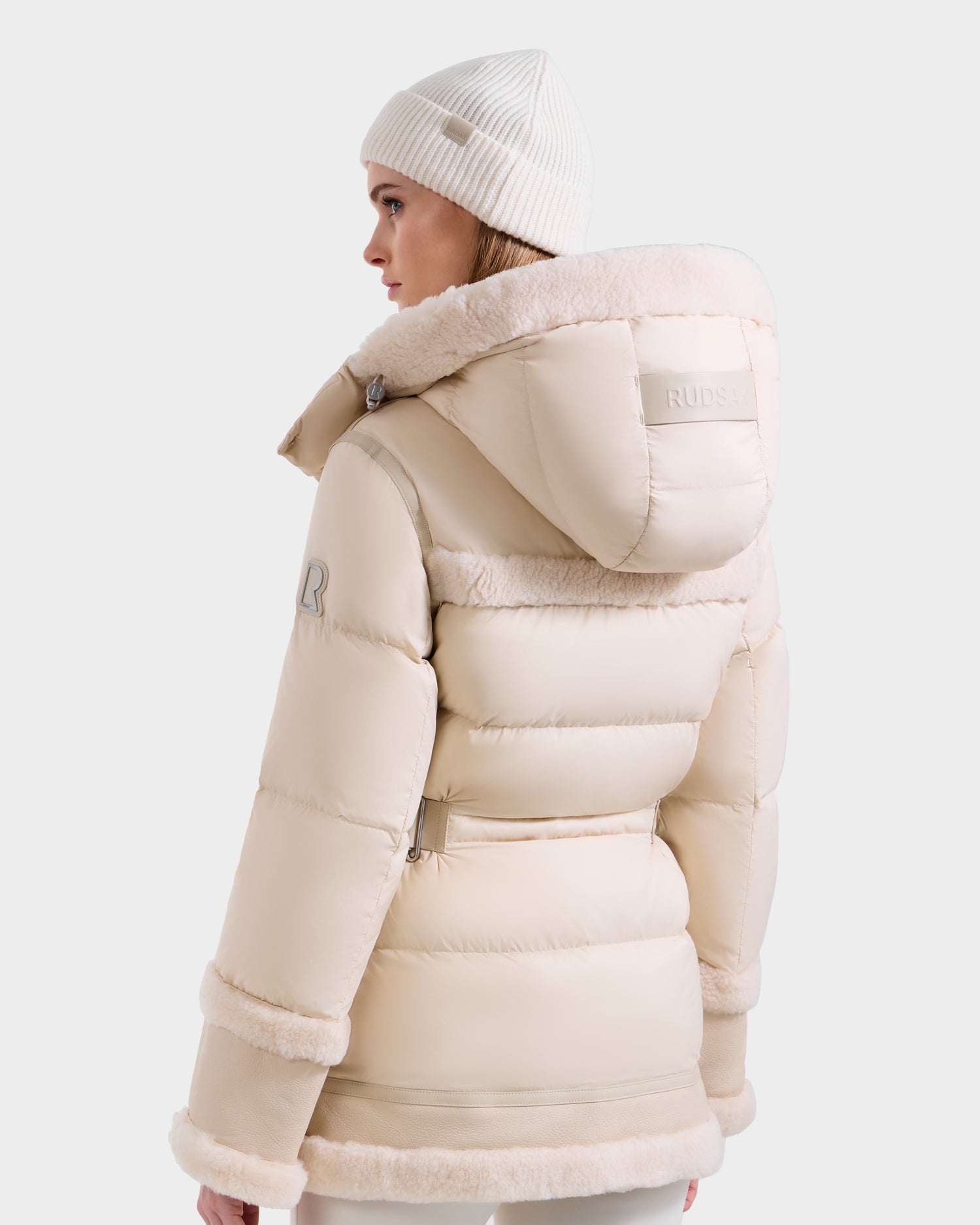 A woman models the Rudsak TESSA - 8125615 Vanilla down puffer jacket with a hood and shearling trim, paired with a matching knit beanie. She stands against a light background, showing a chic winter essential look.
