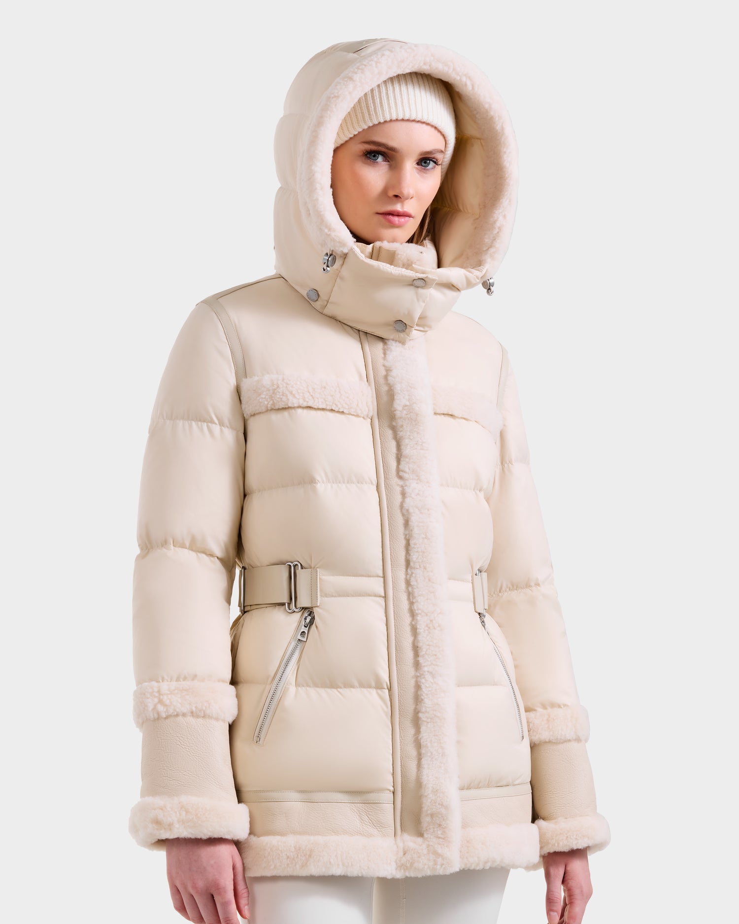 A woman wears the Rudsak TESSA - 8125615 Vanilla, a cream-colored down puffer jacket with shearling trim, hood, matching knit hat, and side pockets—an essential winter piece—against a plain light background.