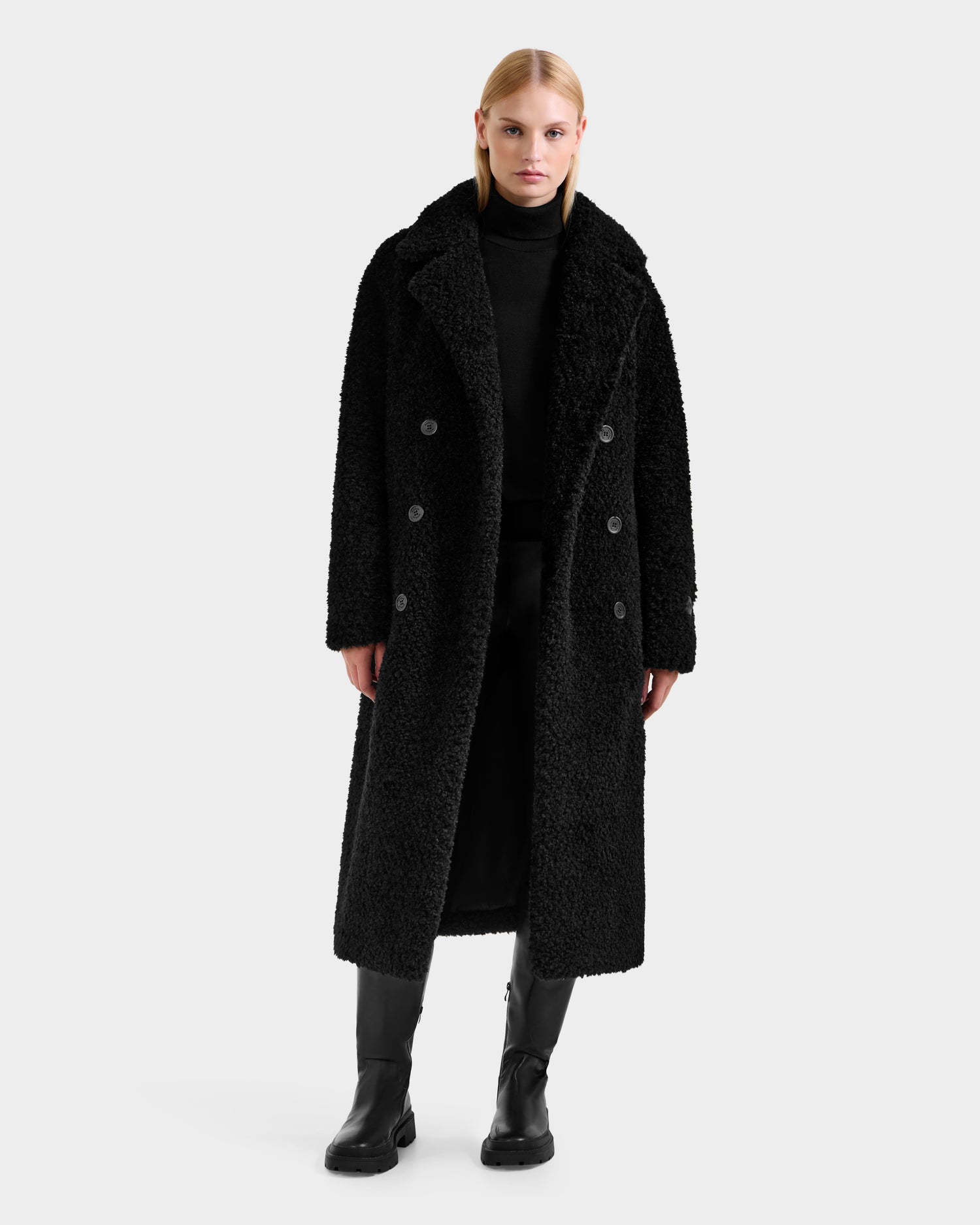 A person stands facing forward against a plain white background, wearing Rudsak’s TINA - 8125968 Black teddy trench coat over a black turtleneck, black pants, and black boots.