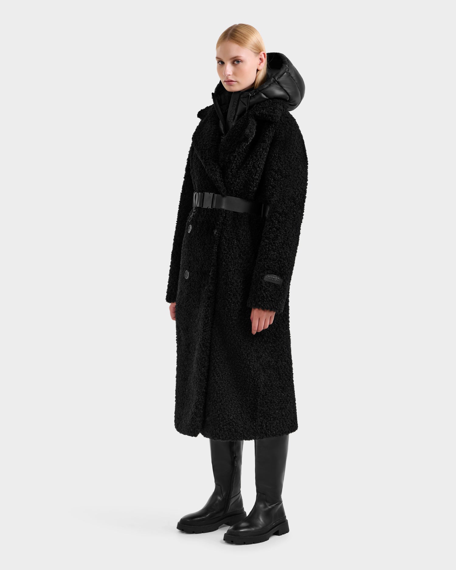 A woman with blonde hair pulled back and a neutral expression wears the Rudsak TINA - 8125968 Black textured teddy trench coat with a belt, styled with black pants and knee-high boots against a plain background.