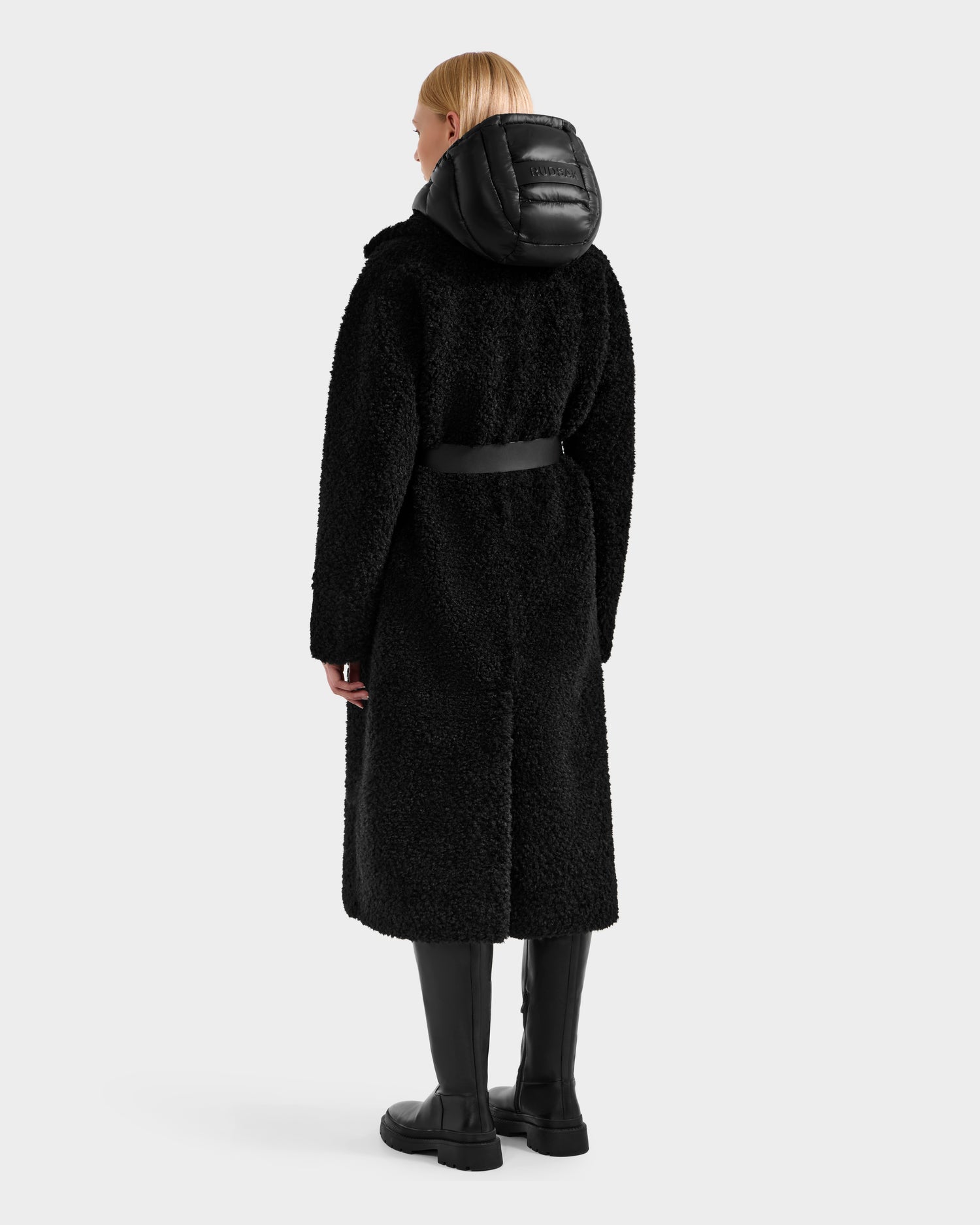 A person with straight blond hair wears the Rudsak TINA - 8125968 Black teddy trench coat with a puffy hood and black belt, paired with black knee-high boots, standing facing away against a plain light background.