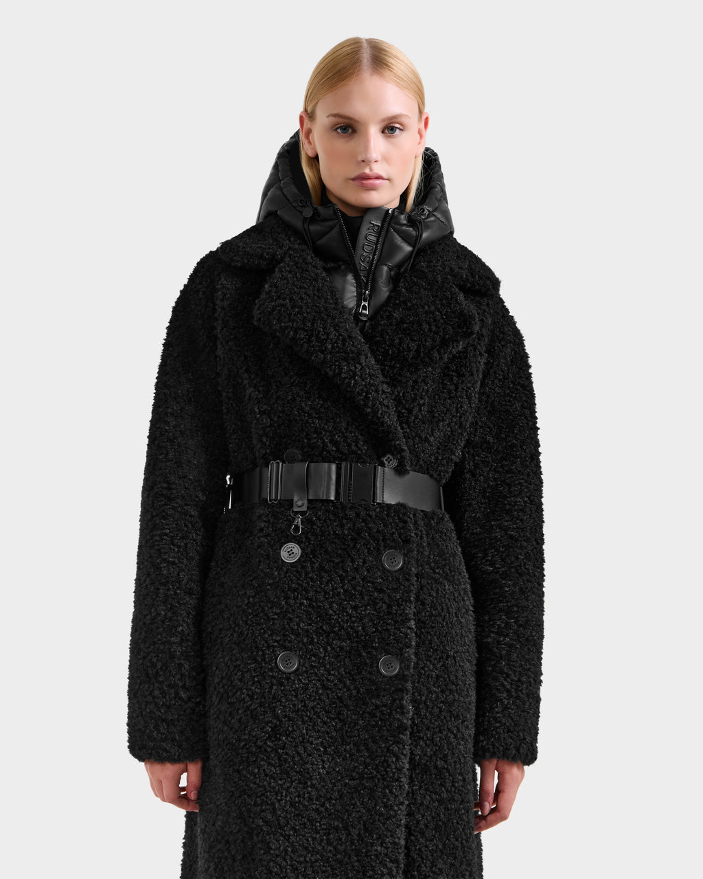 A woman with straight blonde hair models the Rudsak TINA - 8125968 Black trench coat, featuring a textured finish, wide collar, belted waist, and a shiny removable hooded bib, against a plain light background.