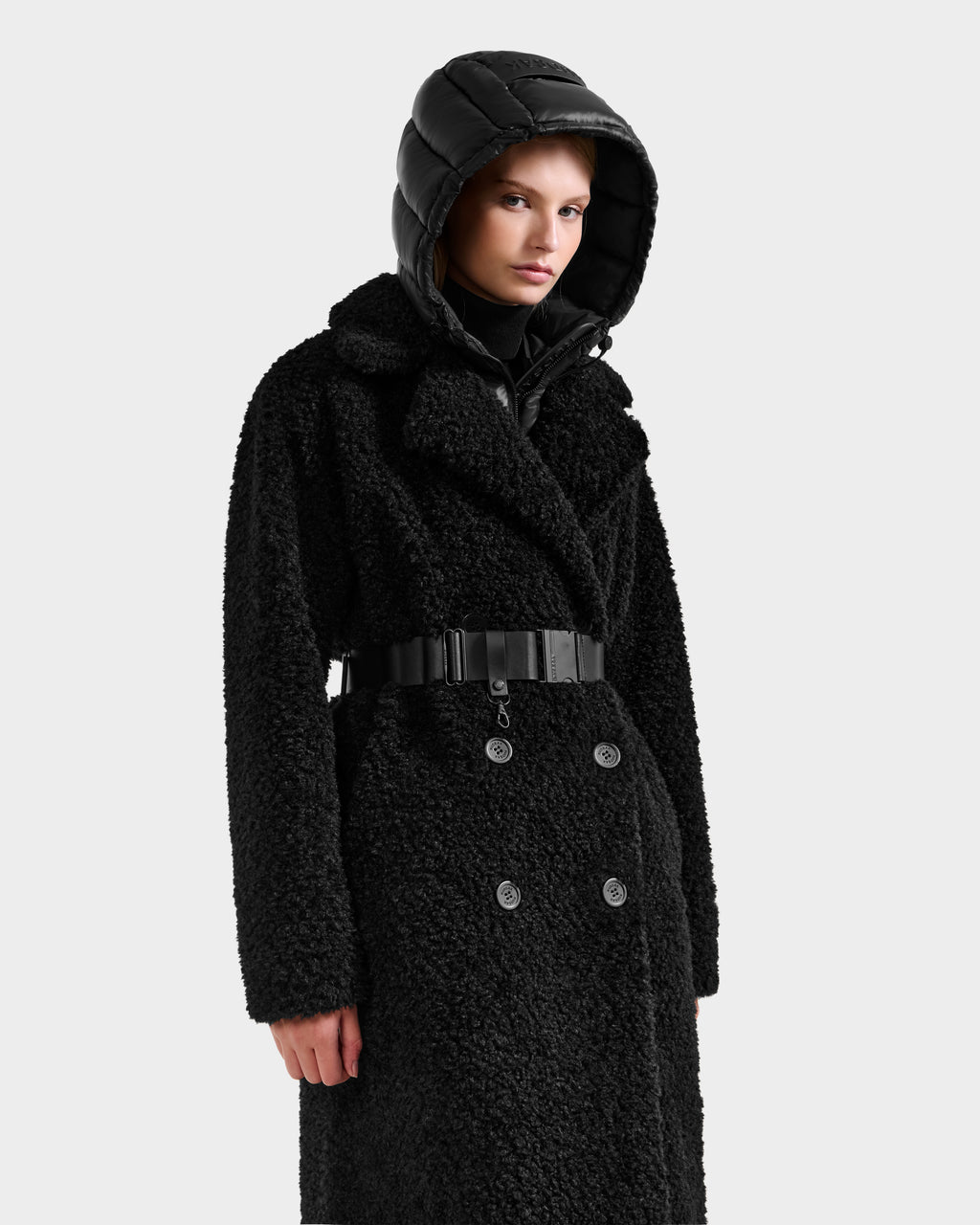 A person wearing the Rudsak TINA - 8125968 Black textured double-breasted coat with a belt and removable hooded bib stands against a plain white background.