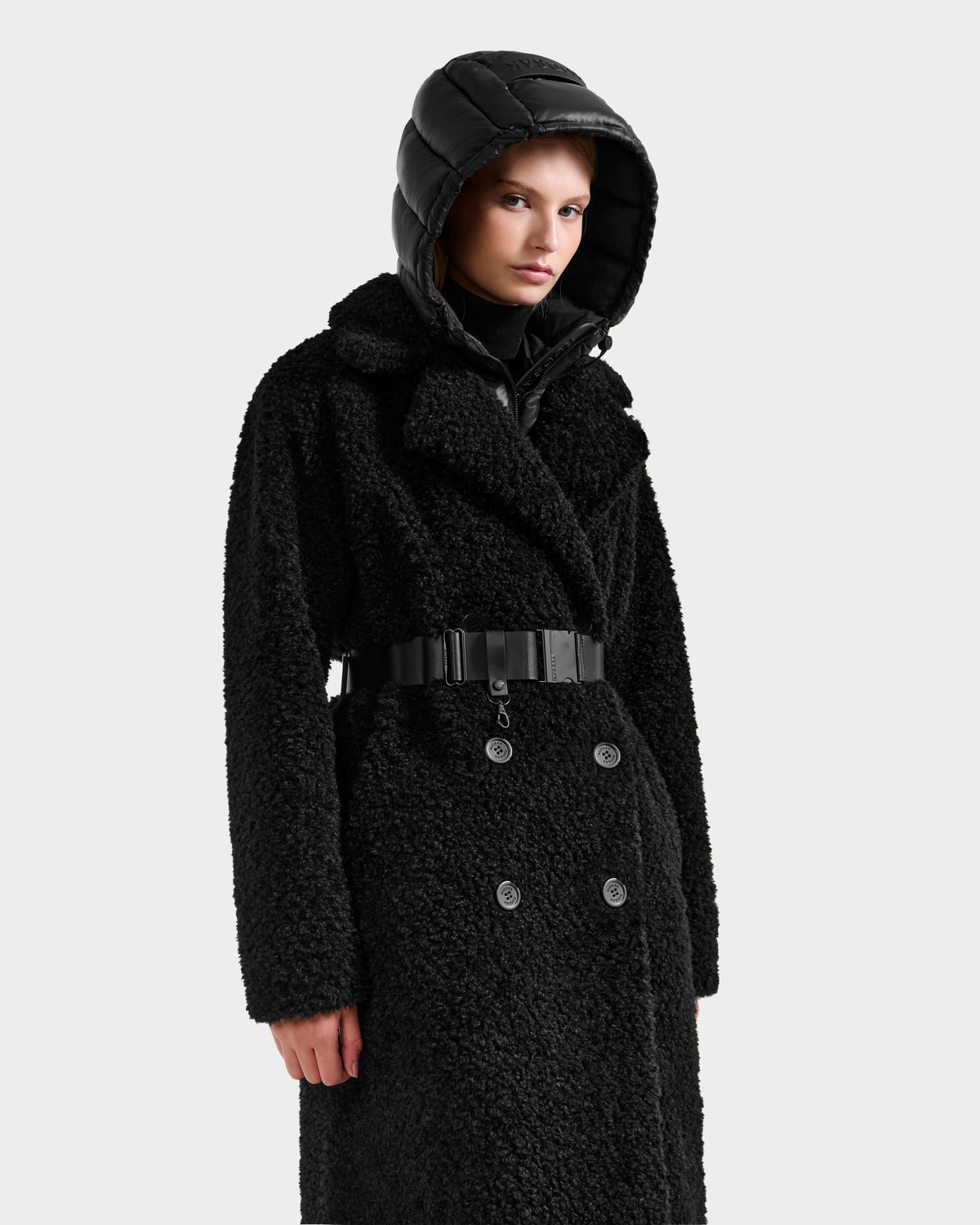 A person wearing the Rudsak TINA - 8125968 Black textured double-breasted coat with a belt and removable hooded bib stands against a plain white background.