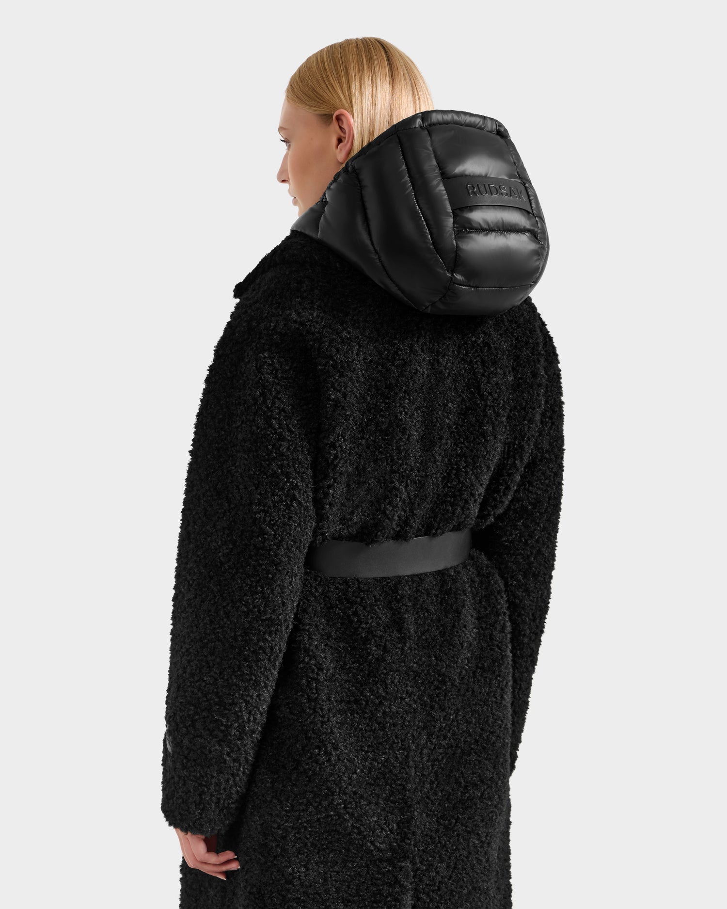 A person with straight blonde hair faces away, wearing the Rudsak TINA - 8125968 Black teddy trench coat with a belted waist and shiny removable hooded bib. The background is light gray.