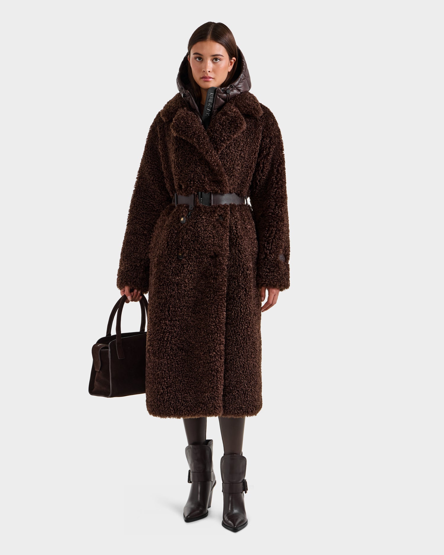 A woman stands facing forward, wearing the Rudsak TINA - 8125968 Espresso faux fur teddy trench coat with a belted waist and dark ankle boots. She holds a matching handbag, her straight brown hair framing her serious expression.