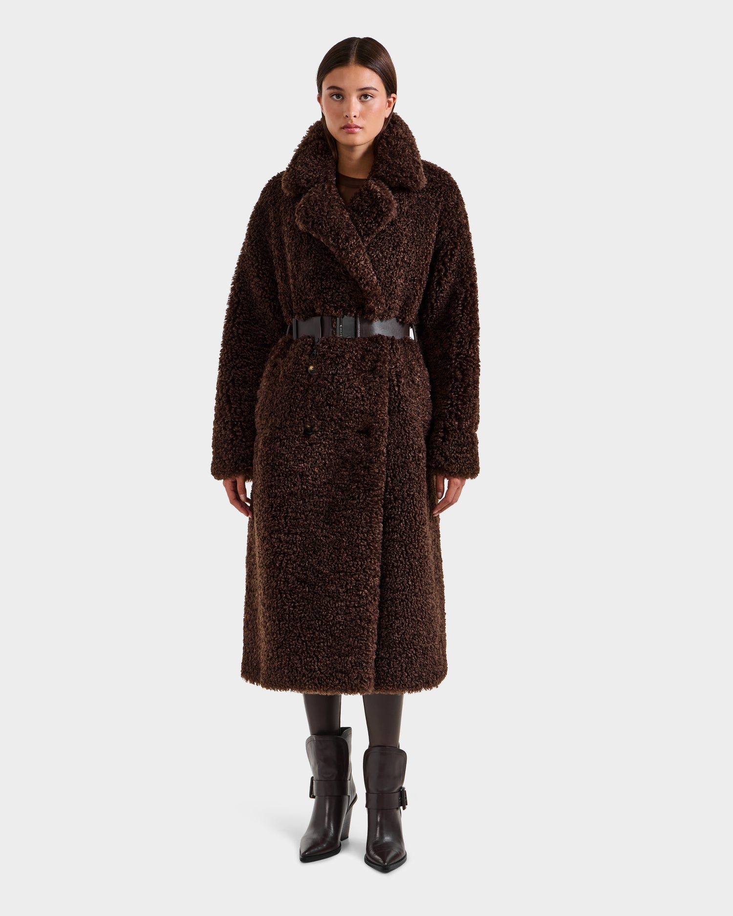 A woman wears the Rudsak TINA - 8125968 Espresso, a long dark brown faux fur teddy trench coat with wide collar and dark belt, styled with dark leggings and black ankle boots, standing against a plain white background.
