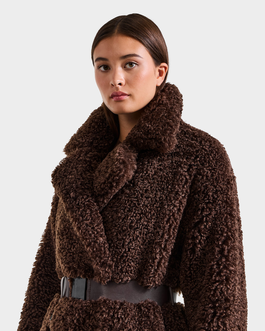 A woman with straight brown hair models the Rudsak TINA - 8125968 Espresso, a dark brown faux fur teddy trench coat with a wide collar and black belt, posed against a simple light background.