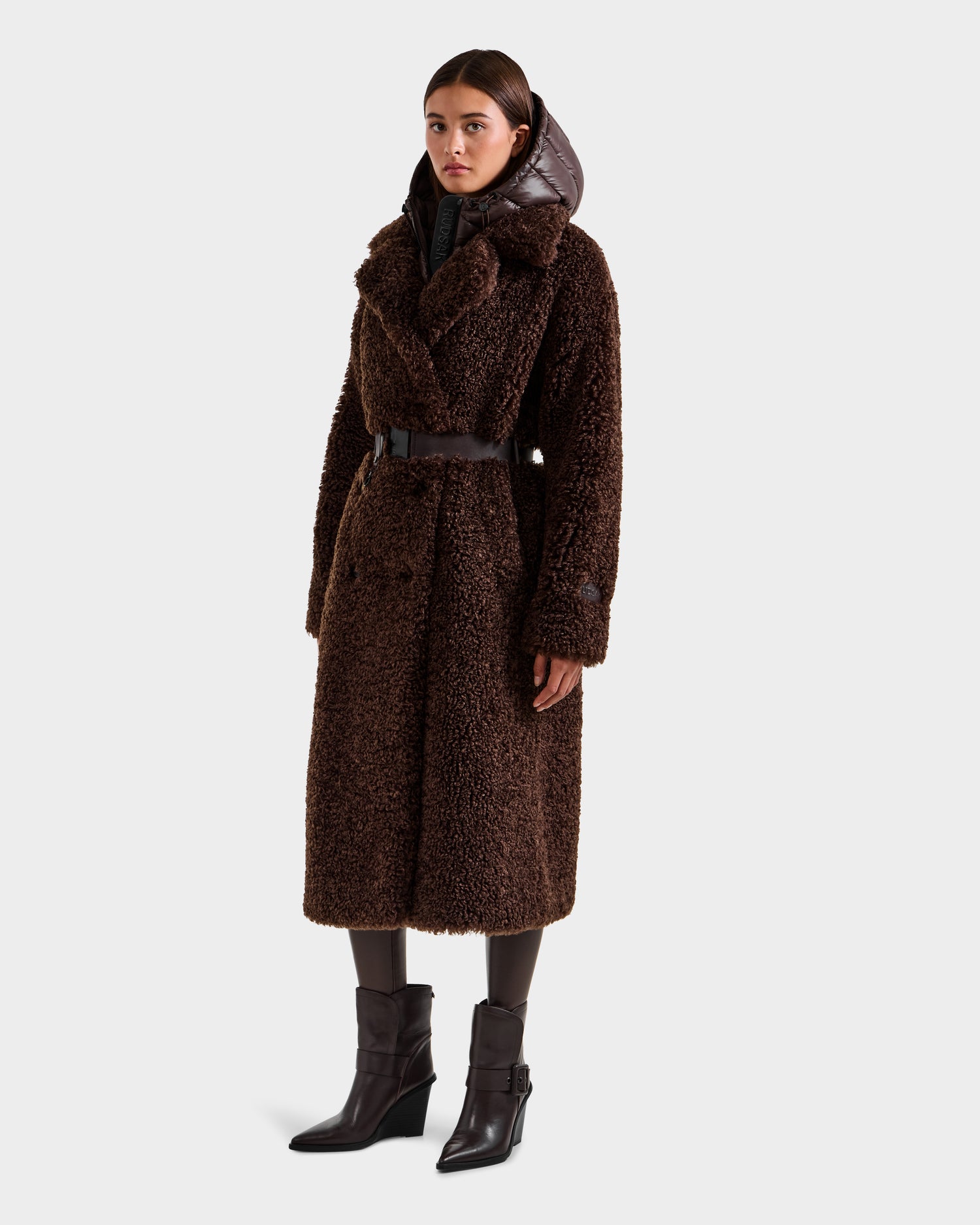 A woman wears the Rudsak TINA - 8125968 Espresso long faux fur teddy trench coat with a black belt, layered over a black hooded garment, dark tights, and black heeled ankle boots, standing against a plain light background.