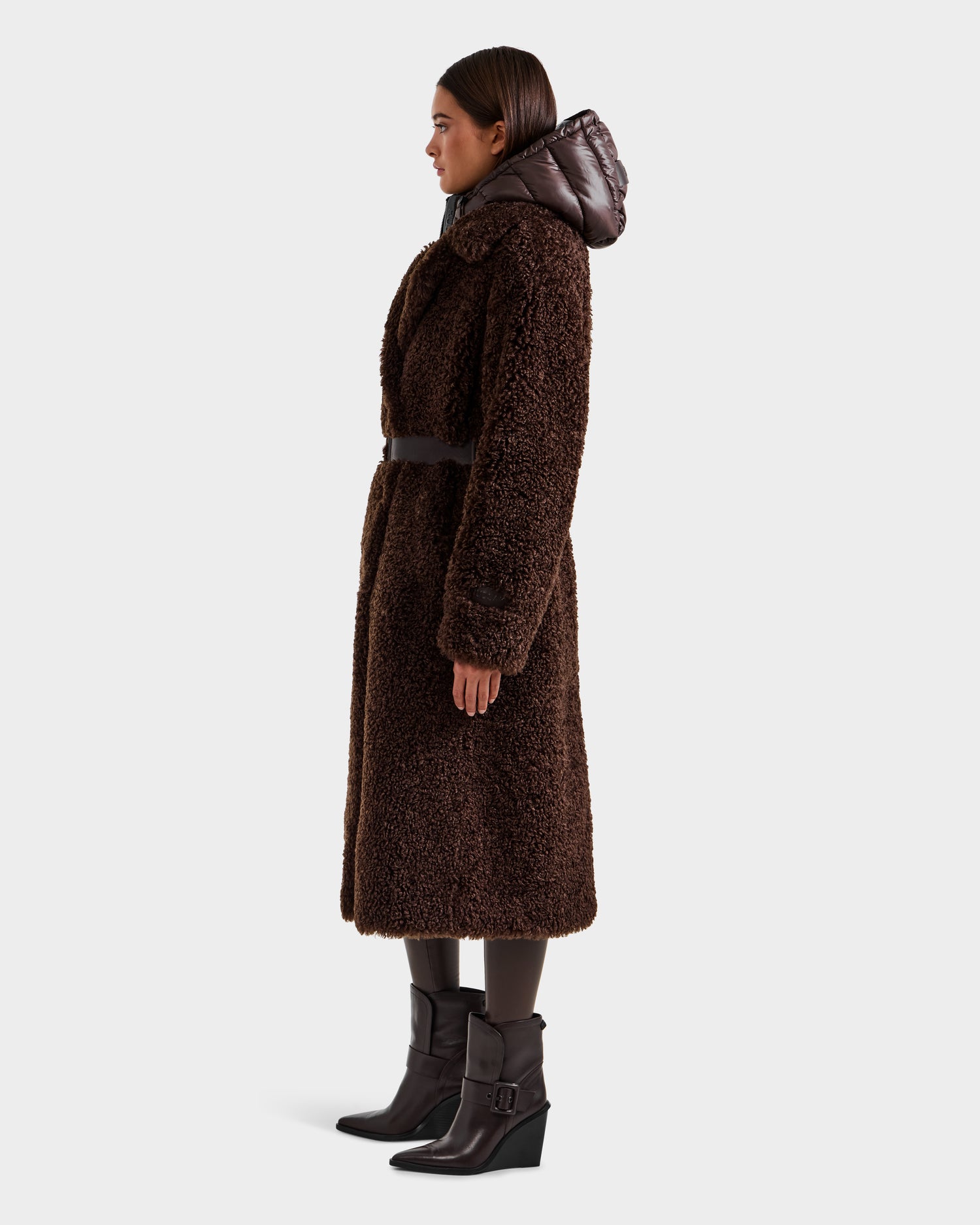A woman in profile wears the Rudsak TINA - 8125968 Espresso long brown faux fur teddy trench coat with a black belt, dark tights, heeled boots, and a removable hooded bib. The background is plain light gray.