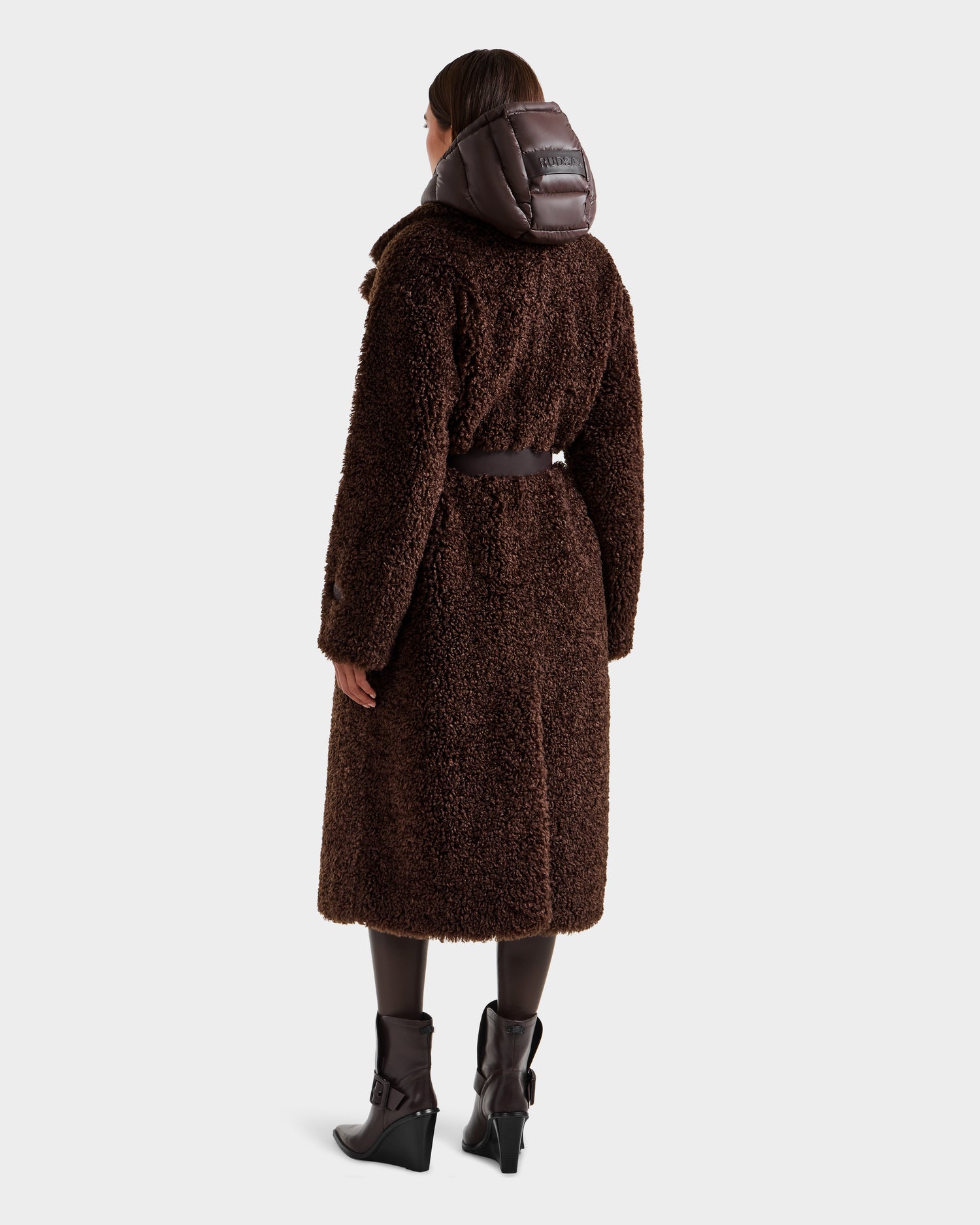 A person faces away, wearing the Rudsak TINA - 8125968 Espresso faux fur teddy trench coat with removable hooded bib. They pair it with dark tights and wedge boots against a plain white background.