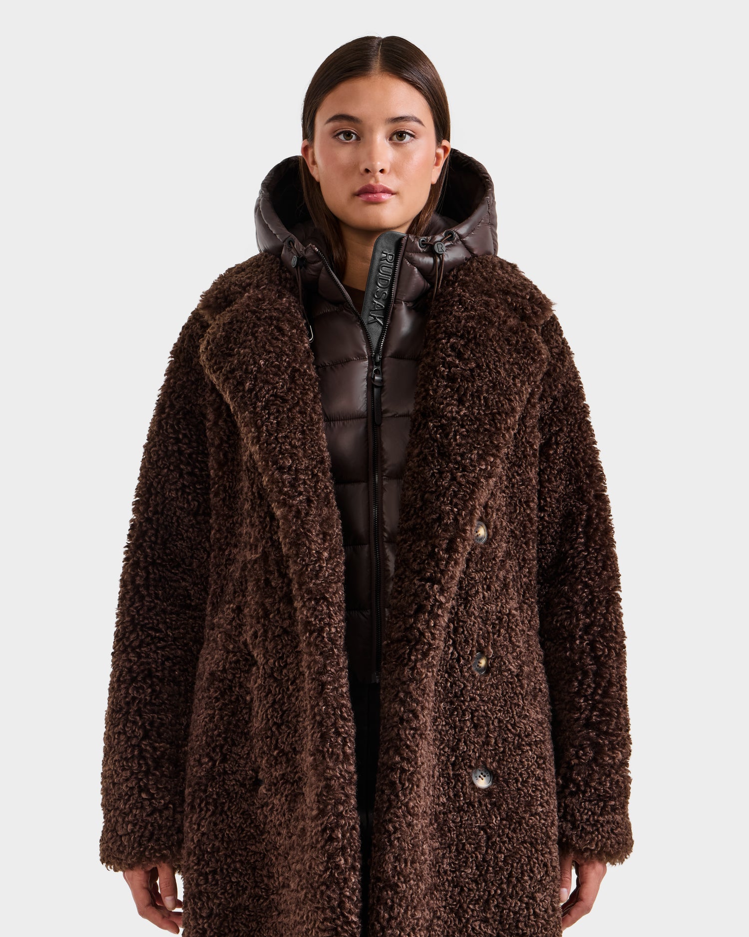 A woman faces forward wearing the Rudsak TINA - 8125968 Espresso dark brown textured faux fur teddy trench coat layered over a shiny puffer jacket with a removable hooded bib. The background is plain white.