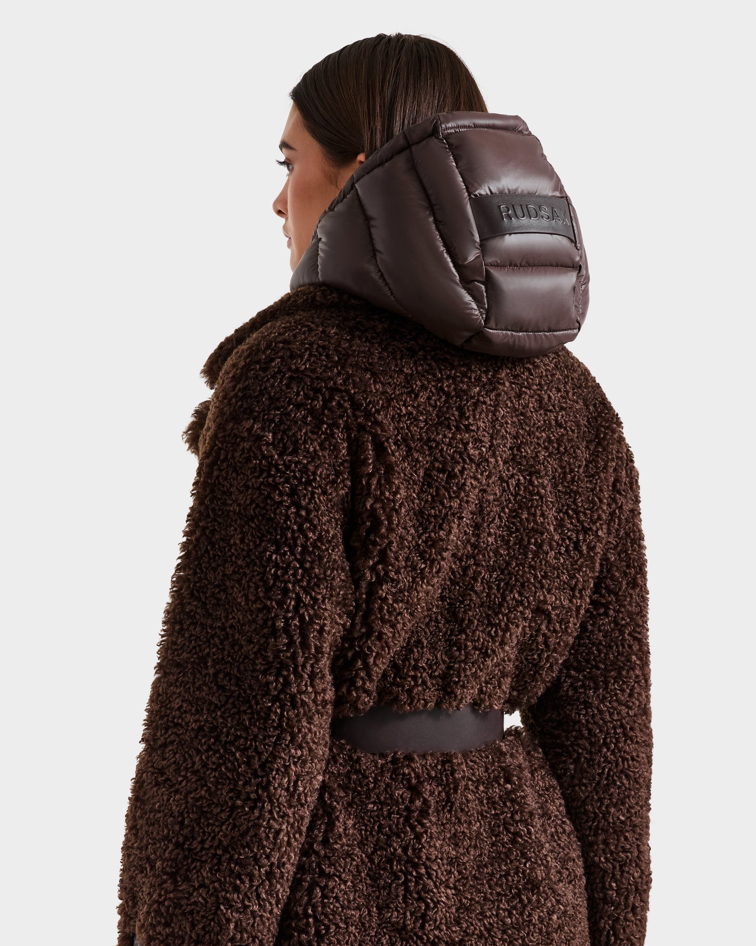 A person wears the Rudsak TINA - 8125968 Espresso women's trench coat in dark brown faux fur with a wide collar, shiny padded removable hooded bib, patch on the back, visible belt, and hood details.