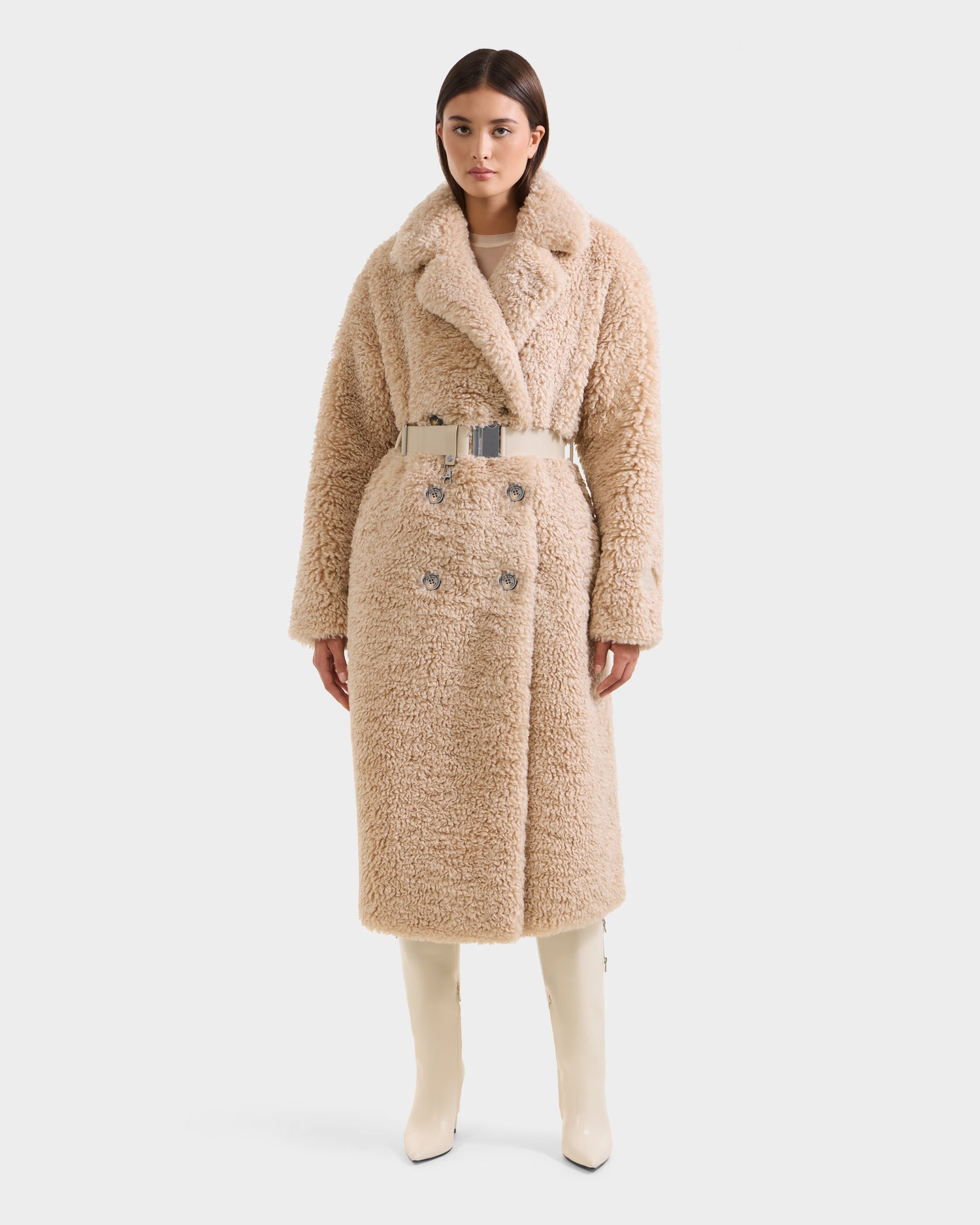 A woman wears the Rudsak TINA - 8125968 Vanilla, a long beige faux fur teddy trench coat with large lapels, double-breasted buttons, and a wide metallic belt. She pairs it with cream knee-high boots, standing against a plain background.