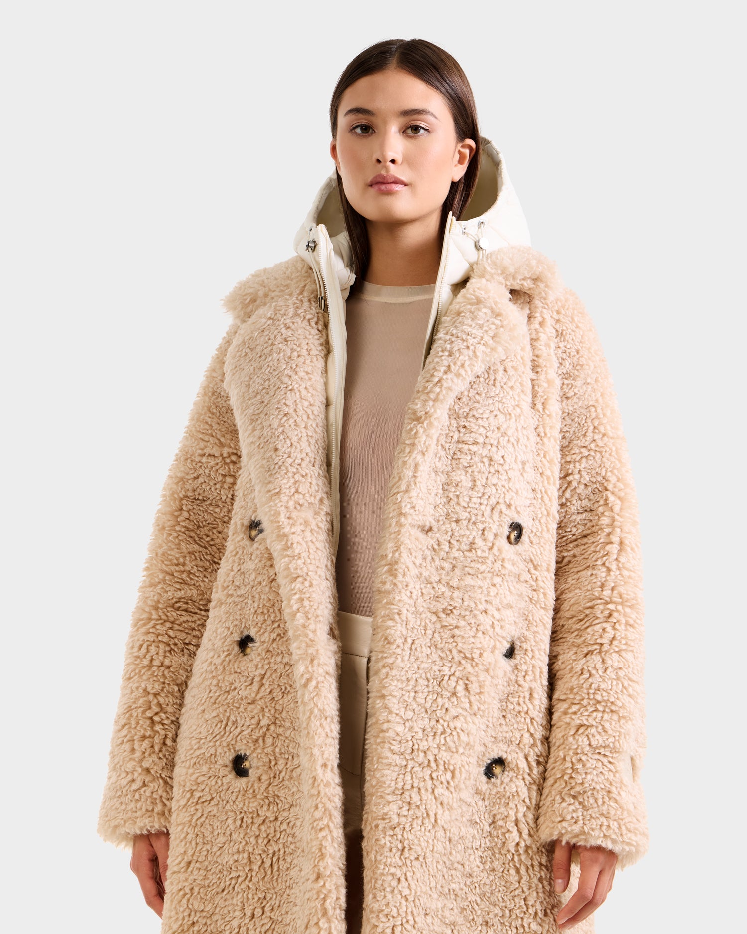 A woman wears the Rudsak TINA - 8125968 Vanilla long, double-breasted faux fur teddy trench coat with removable hooded bib over a light, semi-sheer top and light pants, standing against a plain light grey background.