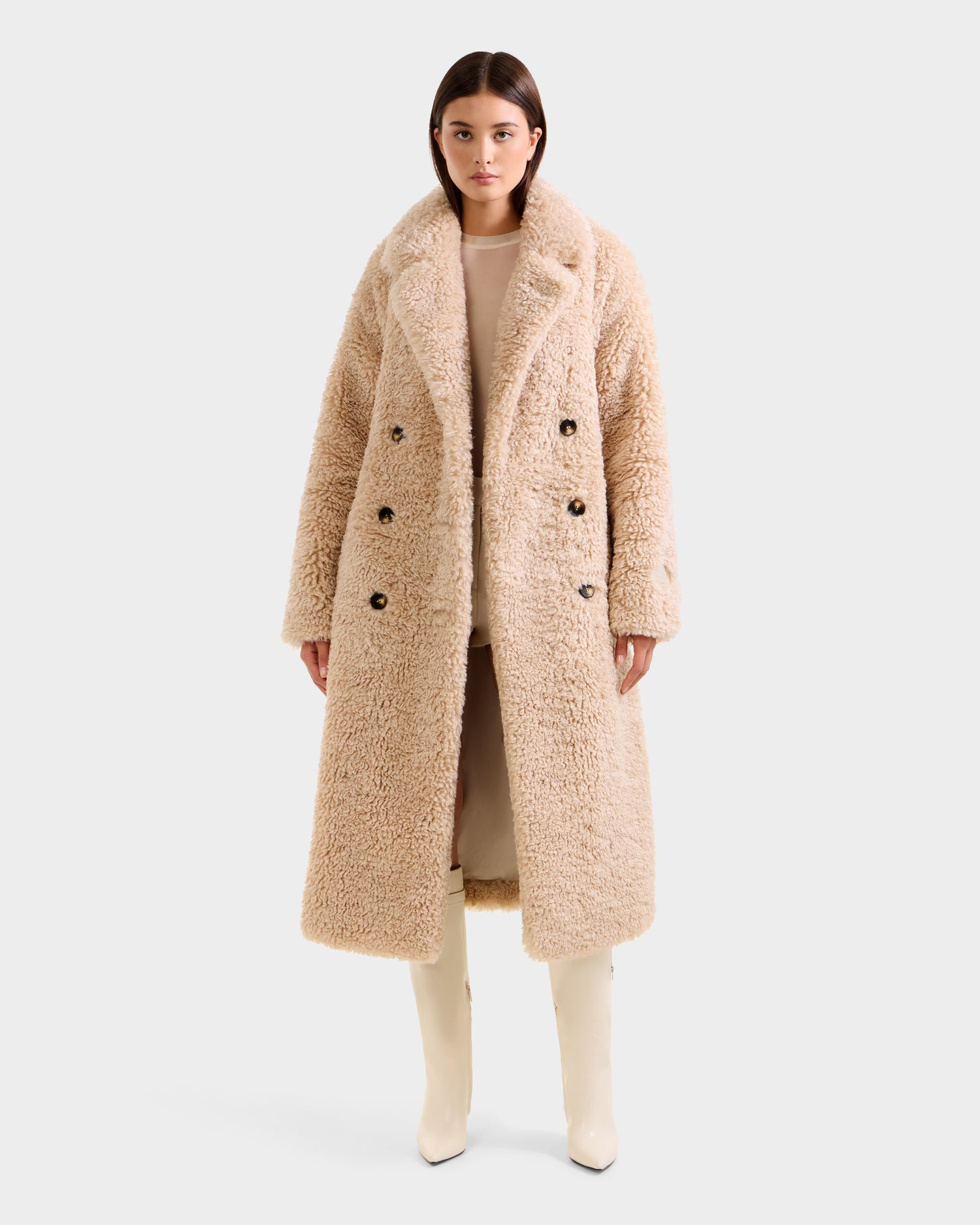 A woman faces forward in the Rudsak TINA - 8125968 Vanilla trench coat, a beige faux fur teddy style with large lapels and a chic double-breasted design, paired with cream knee-high boots against a plain white background.