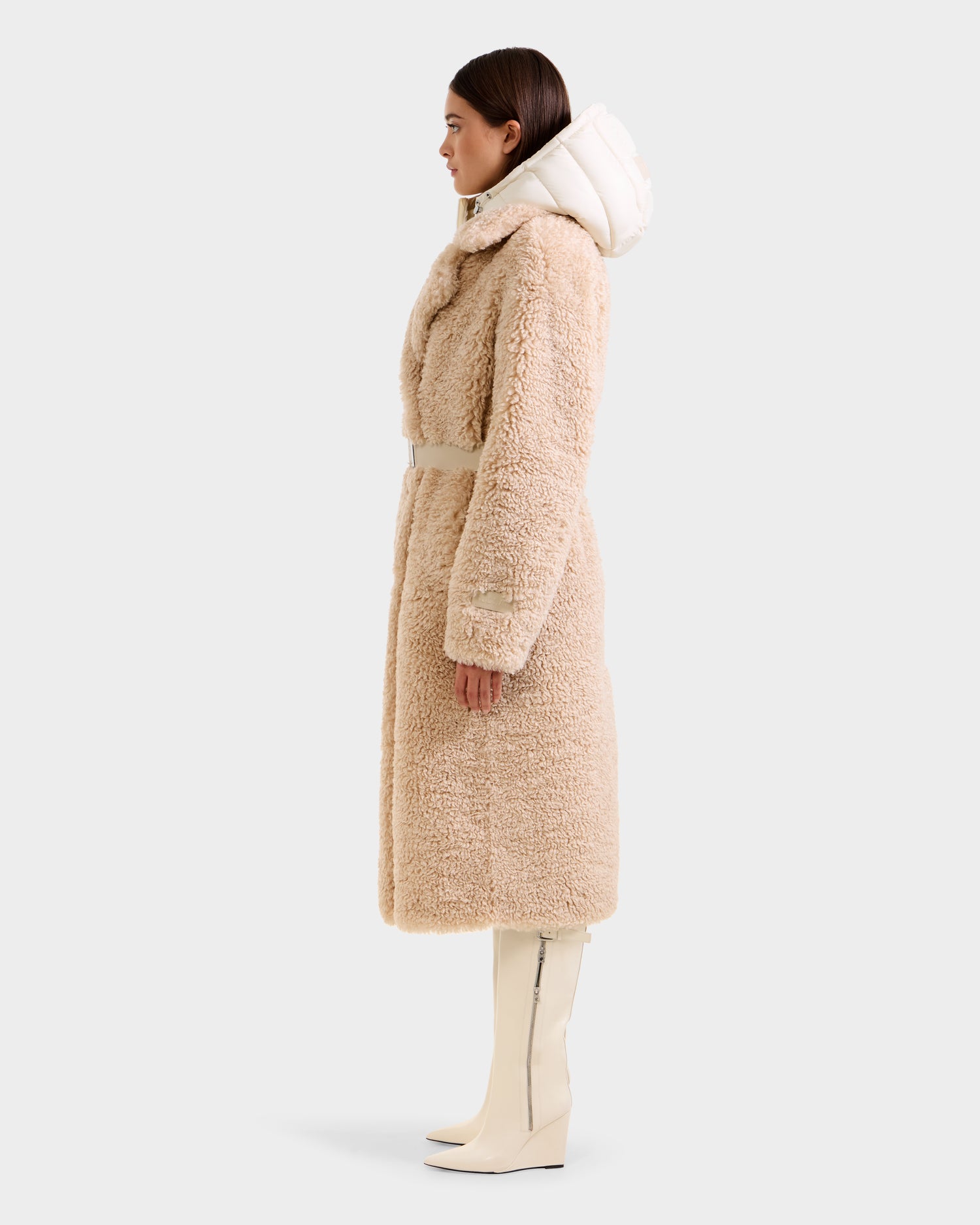A woman in profile wears the Rudsak TINA - 8125968 Vanilla faux fur teddy trench coat with a white hood and belt, paired with white knee-high boots, standing against a plain light gray background.