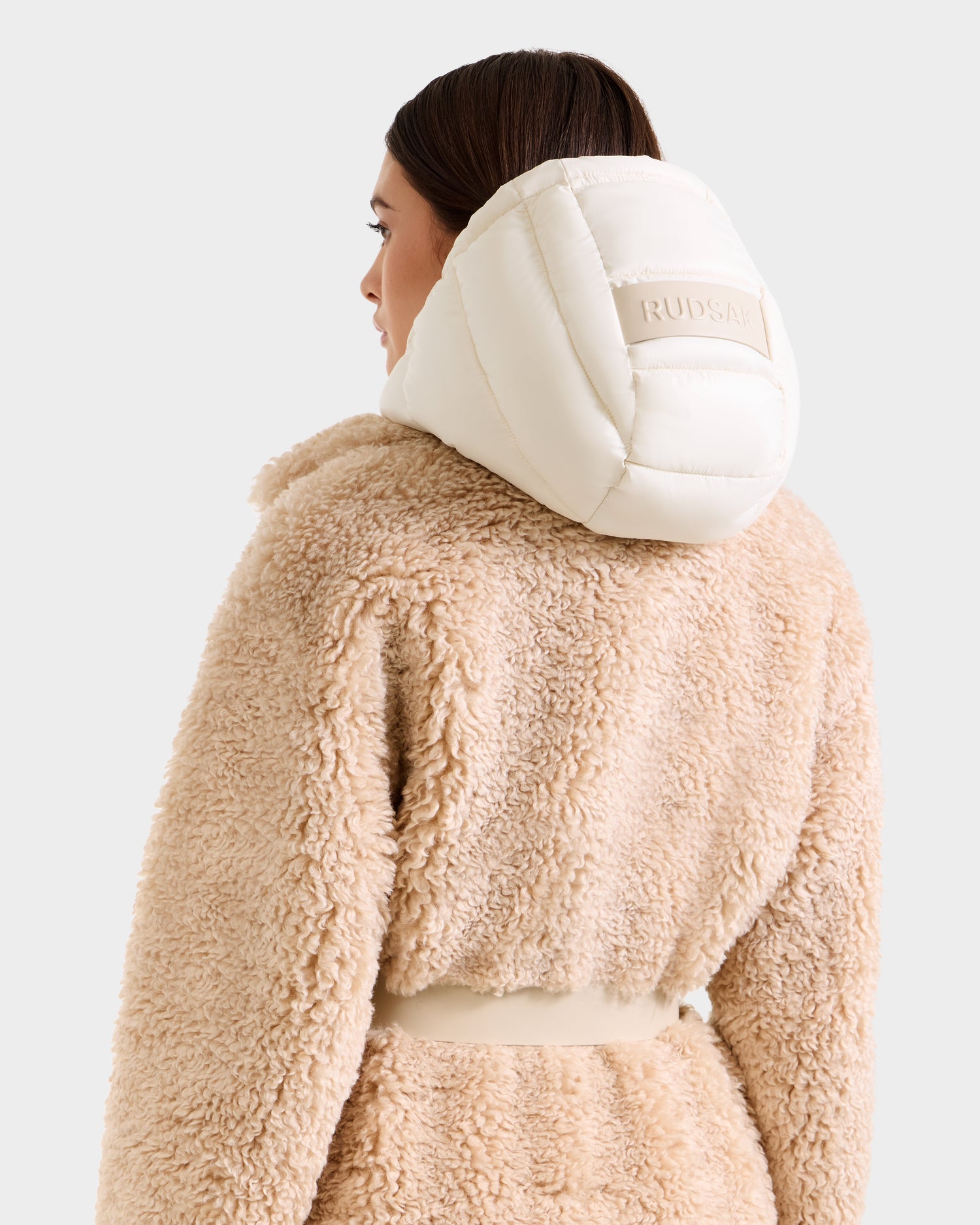 A woman with brown hair wears the Rudsak TINA - 8125968 Vanilla beige faux fur teddy trench coat, featuring a wide collar and padded white hood with "RUDSAK" on the back. She faces away, showcasing the stylish coat’s back and side.