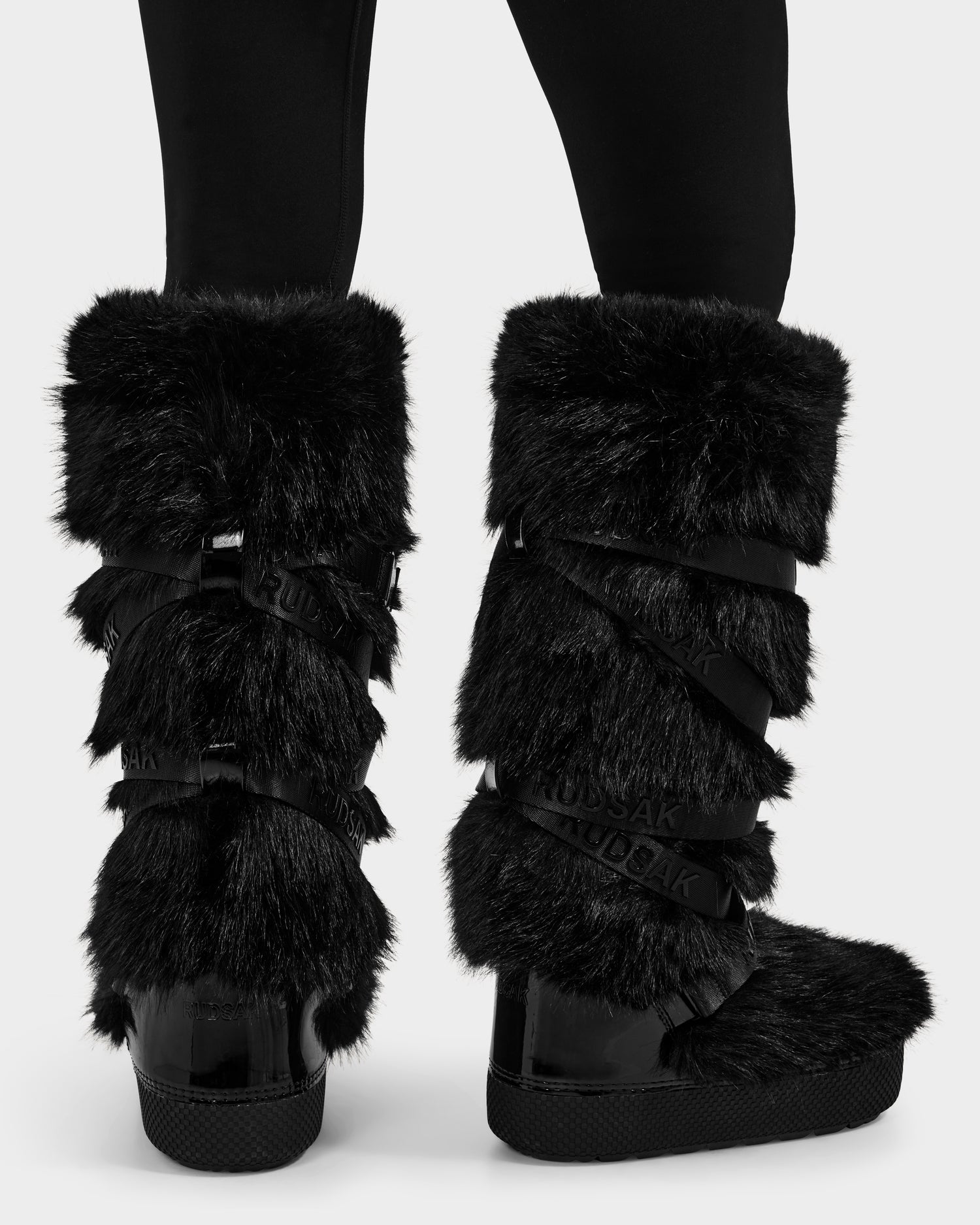 A person wearing Rudsak TORIN - 8225029 Black women's faux fur moon boots with crisscross straps stands on a white background. The fluffy mid-calf après-ski boots are paired with black leggings for a cozy winter look.