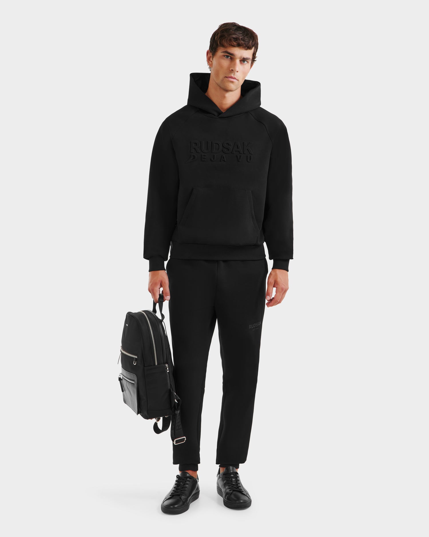 A man stands against a plain background wearing Rudsaks TREVIS - 6525599 Black cotton-blend hoodie with a debossed logo, black jogger pants, and sneakers. He holds a black backpack in his left hand and looks directly at the camera.