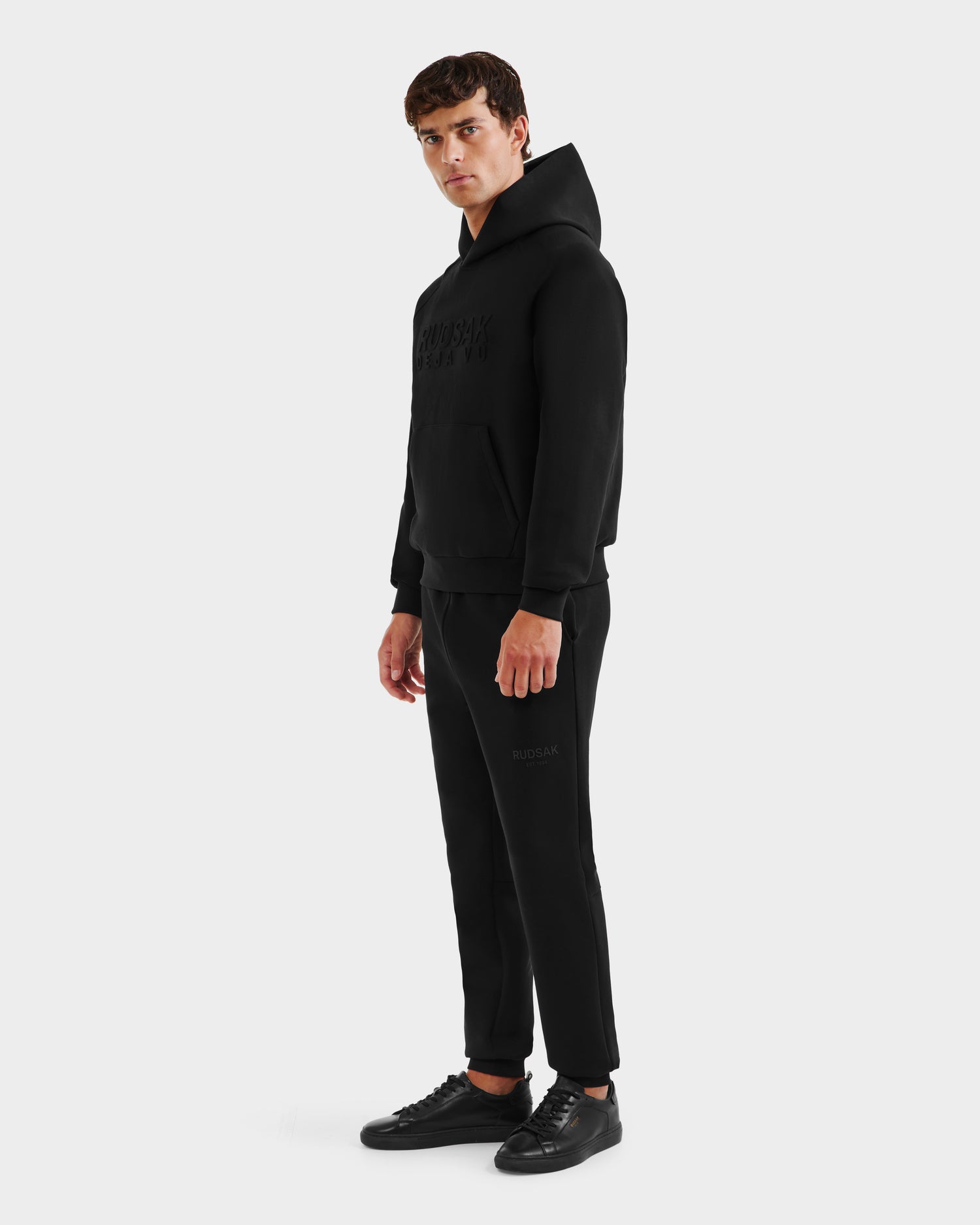 A man stands against a plain background, wearing the Rudsak TREVIS - 6525599 Black men’s cotton-blend hoodie with matching black joggers and sneakers, facing slightly to the side with his hands by his sides.
