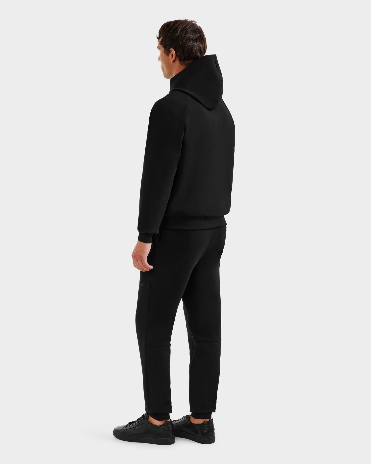 A person faces away, wearing the Rudsak TREVIS - 6525599 Black men’s cotton-blend hoodie with zippered side opening, black sweatpants, and black sneakers against a plain white background.