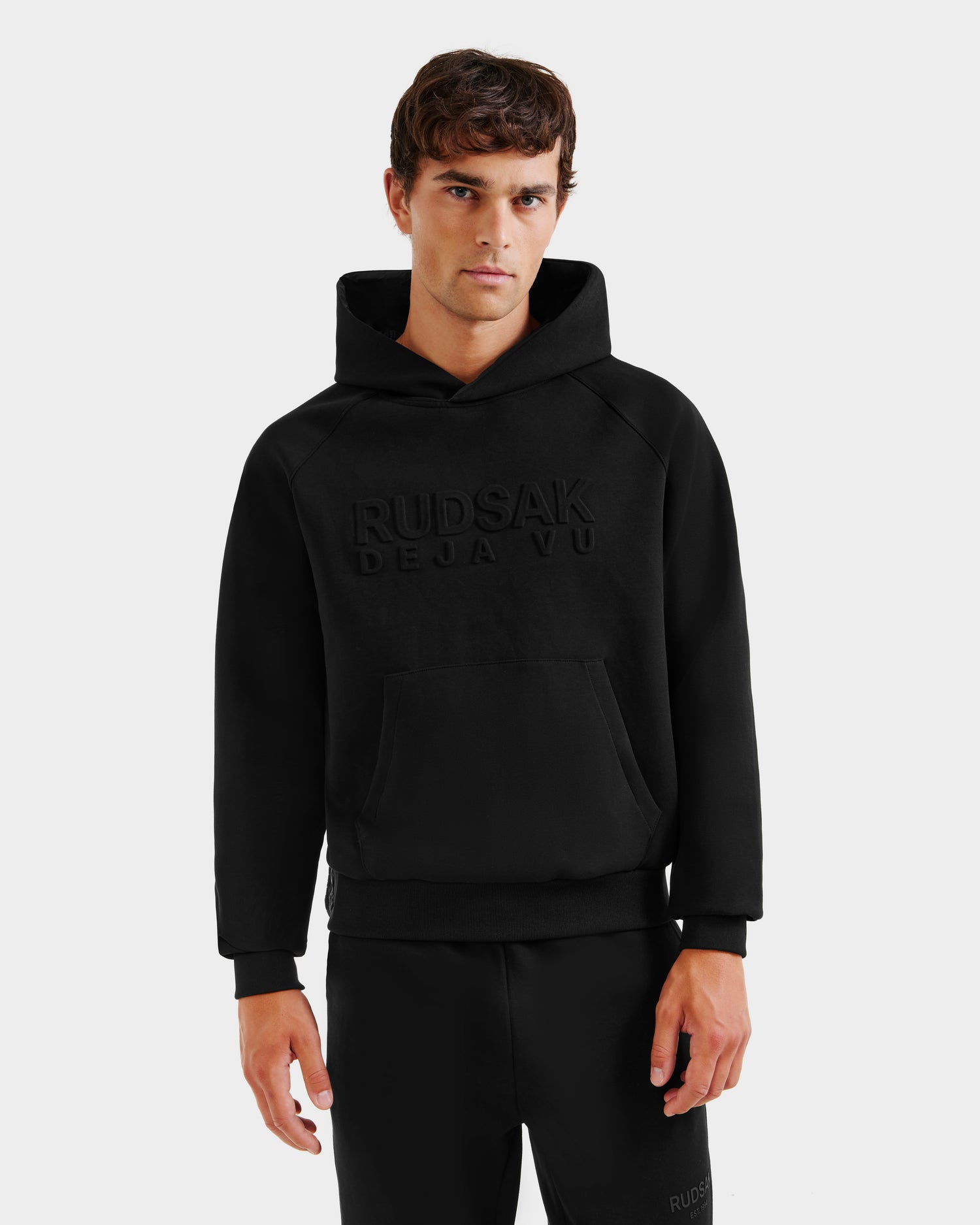 A young man with short brown hair wears the Rudsak TREVIS - 6525599 Black cotton-blend hoodie, featuring an embossed front. He stands against a plain white background, looking directly at the camera with a neutral expression.