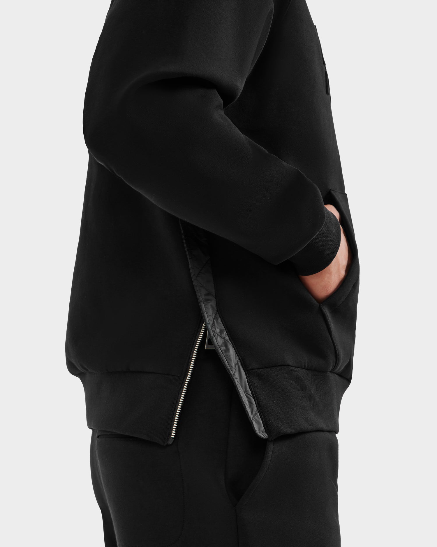A person wearing the Rudsak TREVIS - 6525599 Black men’s cotton-blend hoodie and black pants stands side-on, showcasing its clean, minimal style with a zippered side opening and one hand in the pocket.