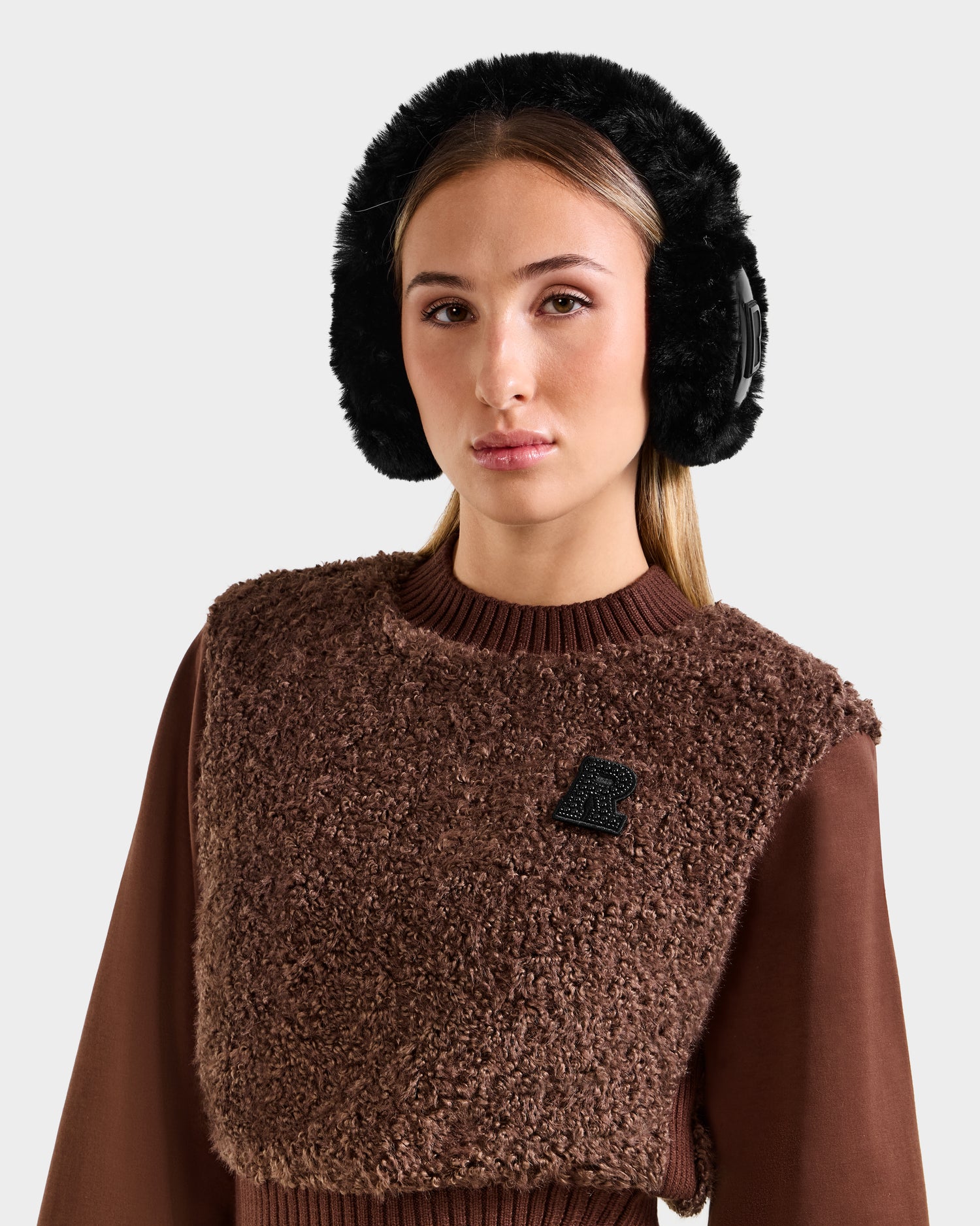 A woman with long, straight hair models the Rudsak VALENCIA - 8425543 Black faux fur earmuffs and a brown textured sweater with a black chest patch, standing against a light gray background.
