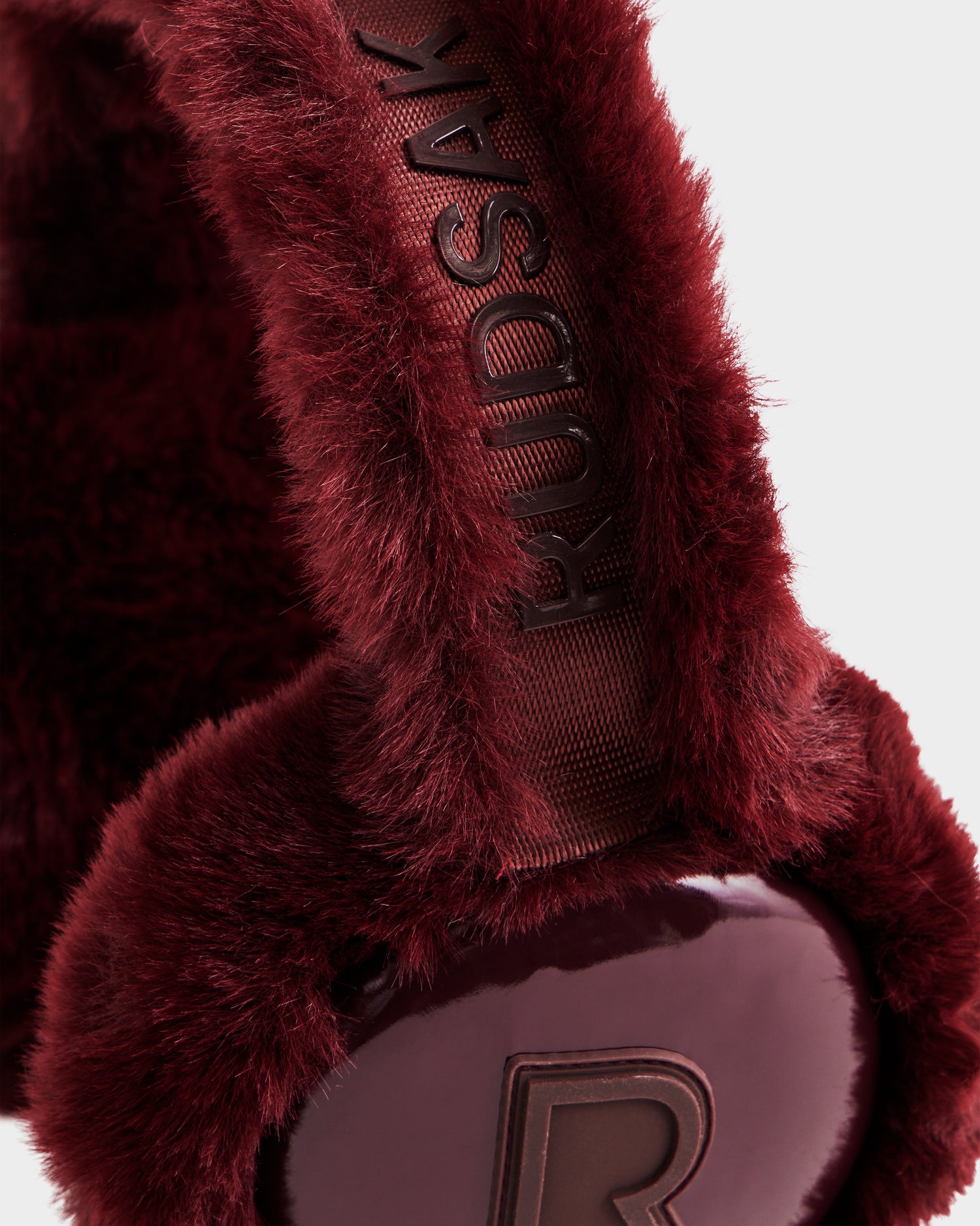 Close-up of VALENCIA - 8425543 Bordeaux earmuffs by Rudsak, made from plush faux fur. The headband features the brand name embossing and a raised “R” on the ear cover—an elegant winter accessory.