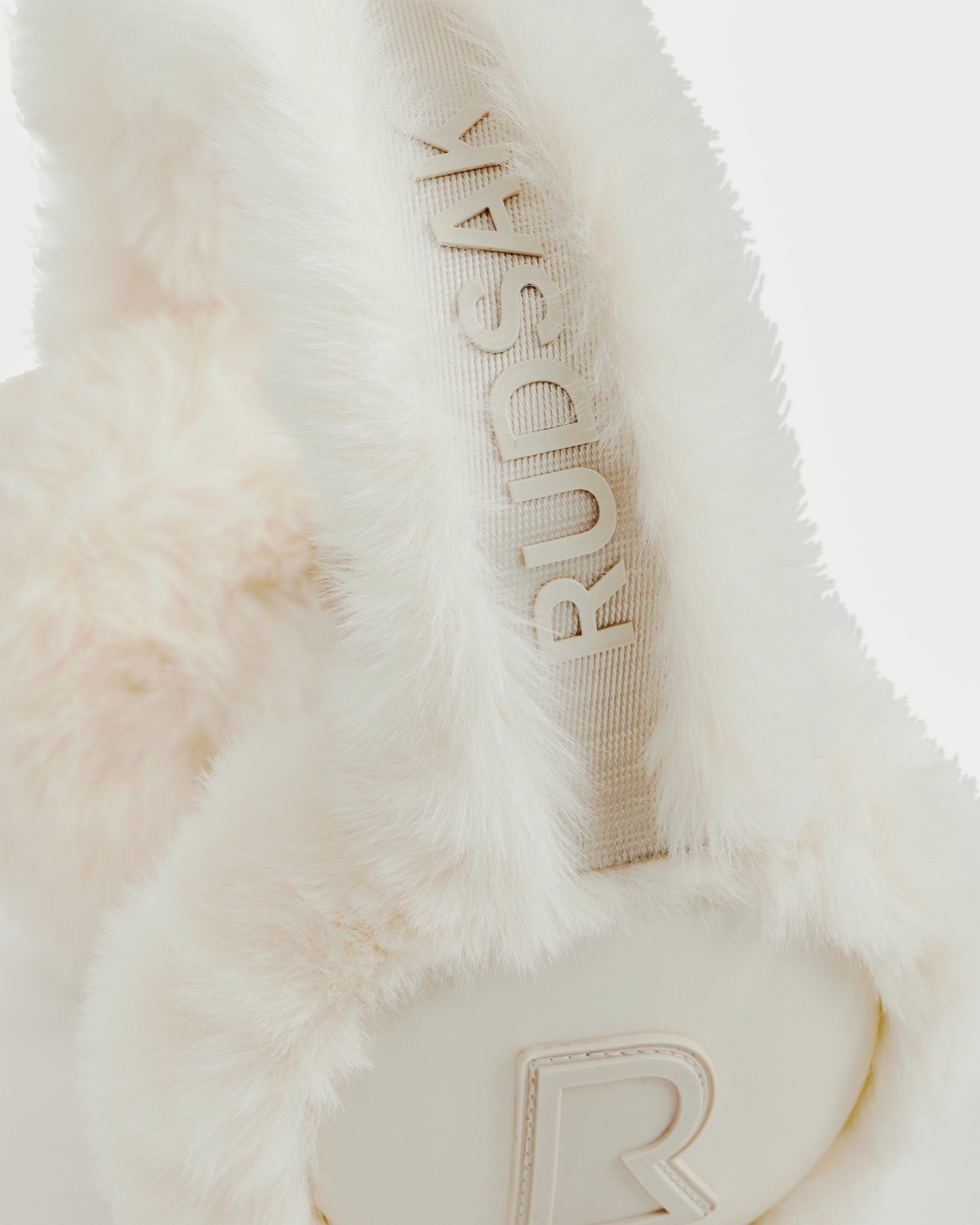 Close-up of white faux fur Rudsak VALENCIA - 8425543 Iceland earmuffs, a stylish winter accessory with the brand name embossed on the headband and a raised R logo on the ear cover, set against a white background.