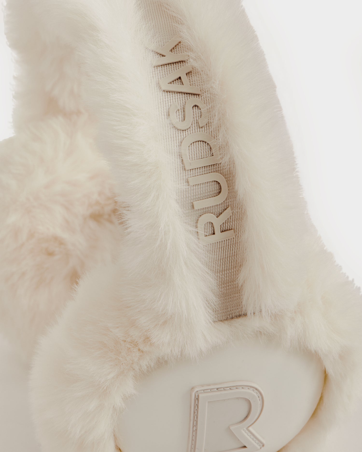 Close-up of the Rudsak VALENCIA - 8425543 Vanilla earmuffs featuring white, fluffy faux fur, an embroidered Rudsak band, and a raised "R" logo—an essential winter accessory.