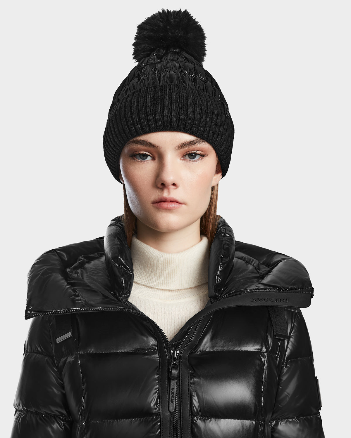 A woman in a black knit pom-pom beanie and the Rudsak VENICE S - L124514 Black knee-length down puffer over a light turtleneck looks straight at the camera with a neutral expression.