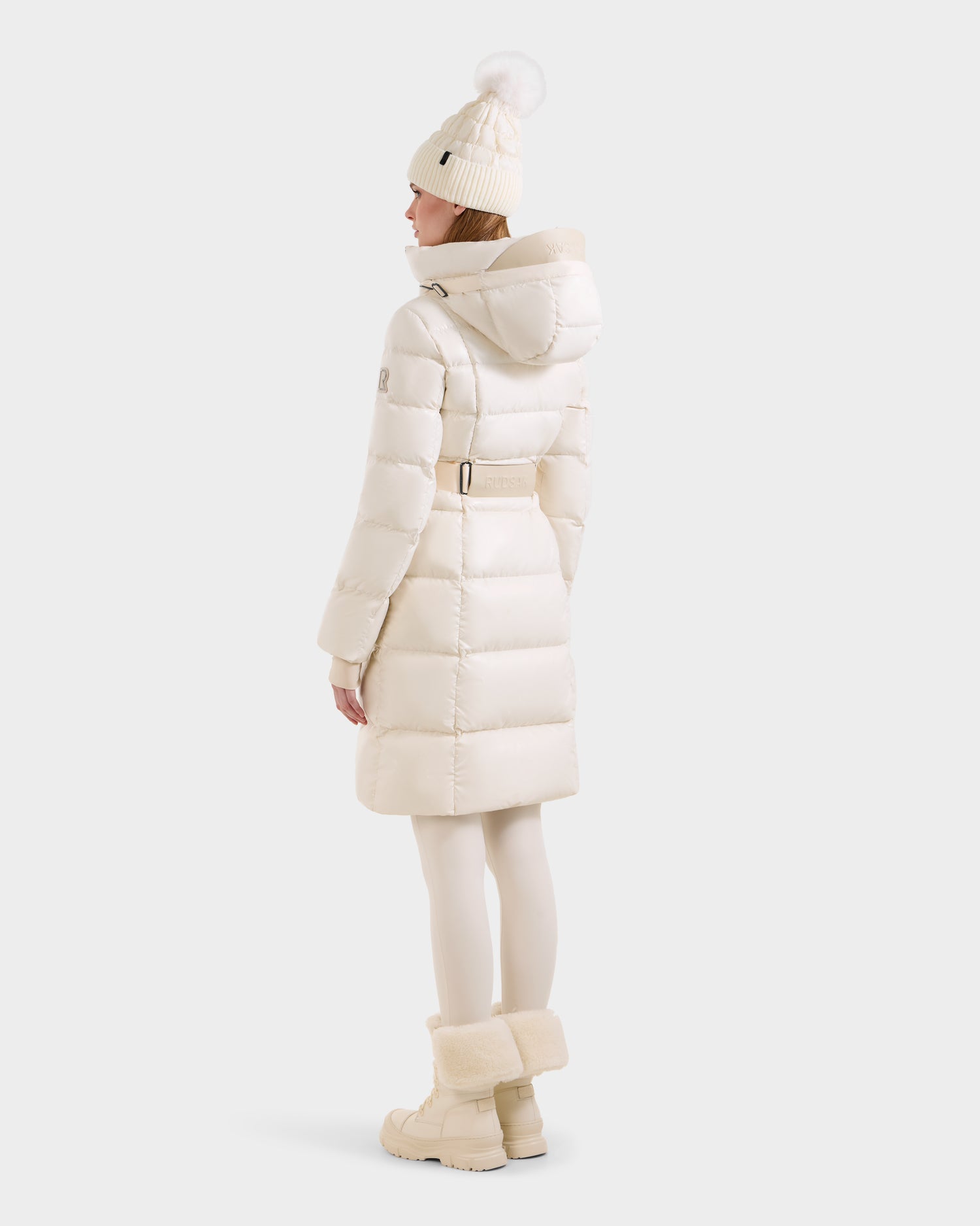 A woman faces away, wearing the Rudsak VENICE S - L124514 Vanilla knee-length belted and hooded puffer, paired with a white pom-pom knit hat, white tights, and beige fur-trimmed boots, against a light gray backdrop.