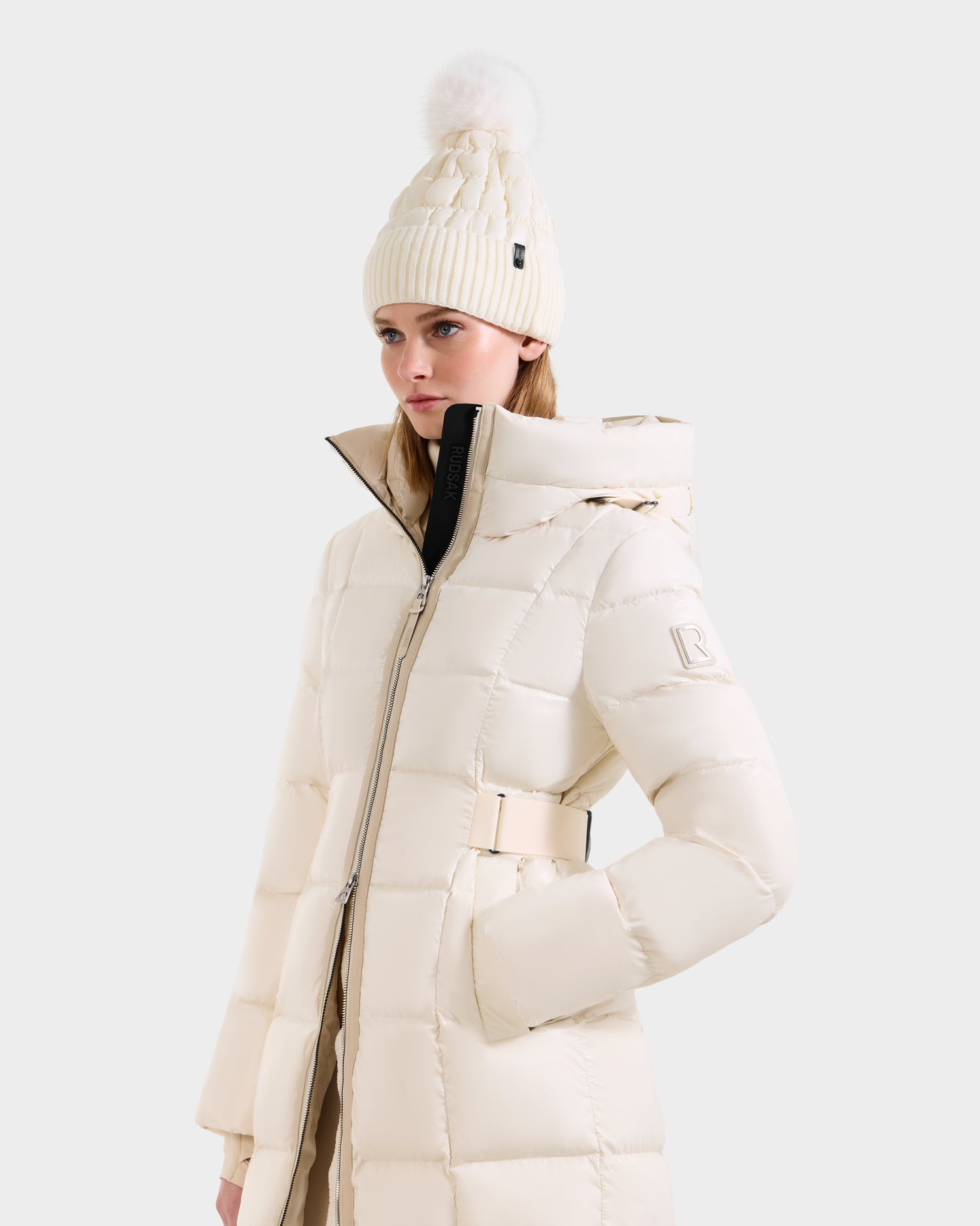 A woman wears the Rudsak VENICE S - L124514 Vanilla cream knee-length down puffer with a hood and a matching pom-pom knit beanie, standing against a plain light background.