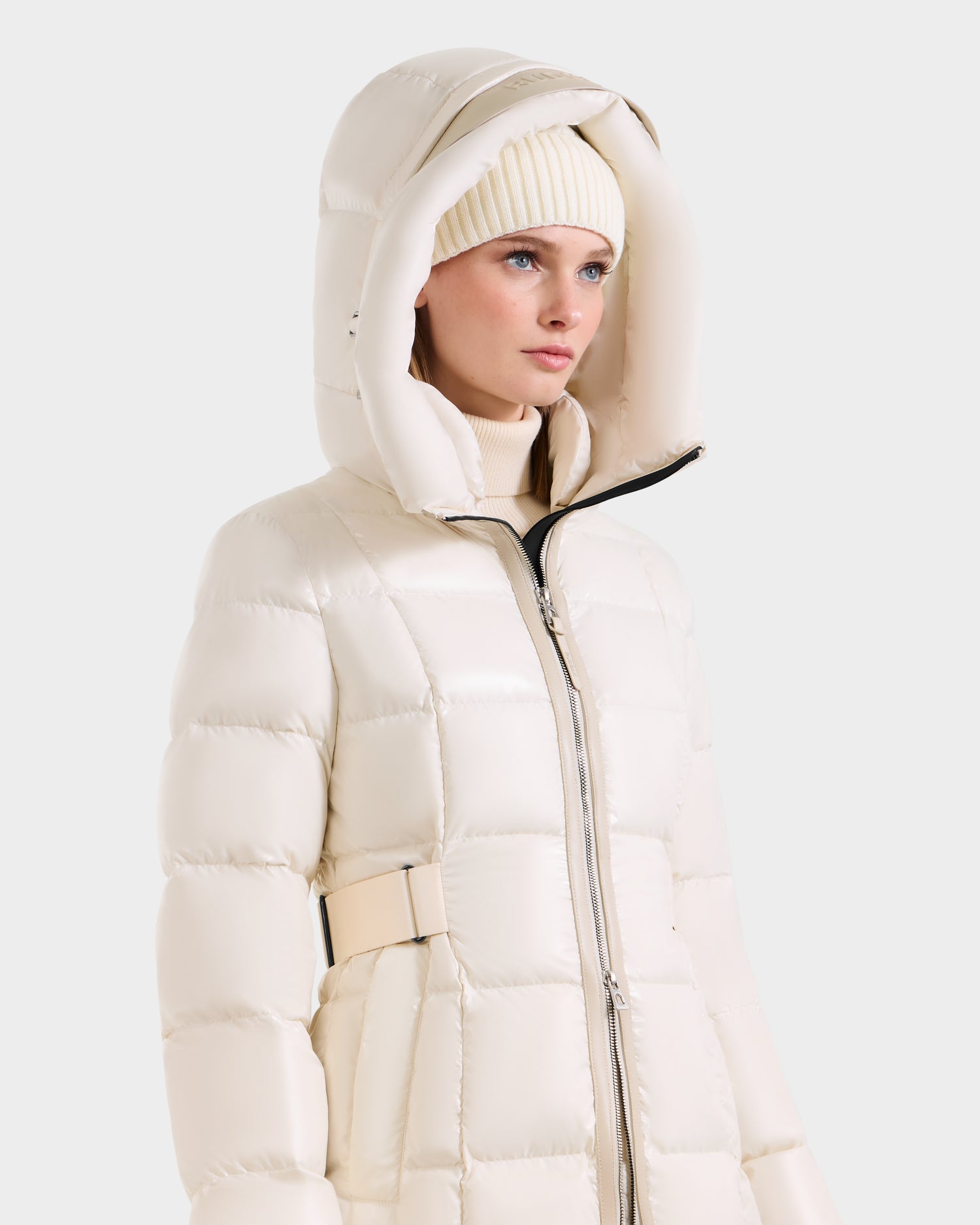 A person wears the Rudsak VENICE S - L124514 Vanilla, a cream knee-length down puffer with hood, paired with a matching knit beanie, looking slightly to the side against a plain light background.