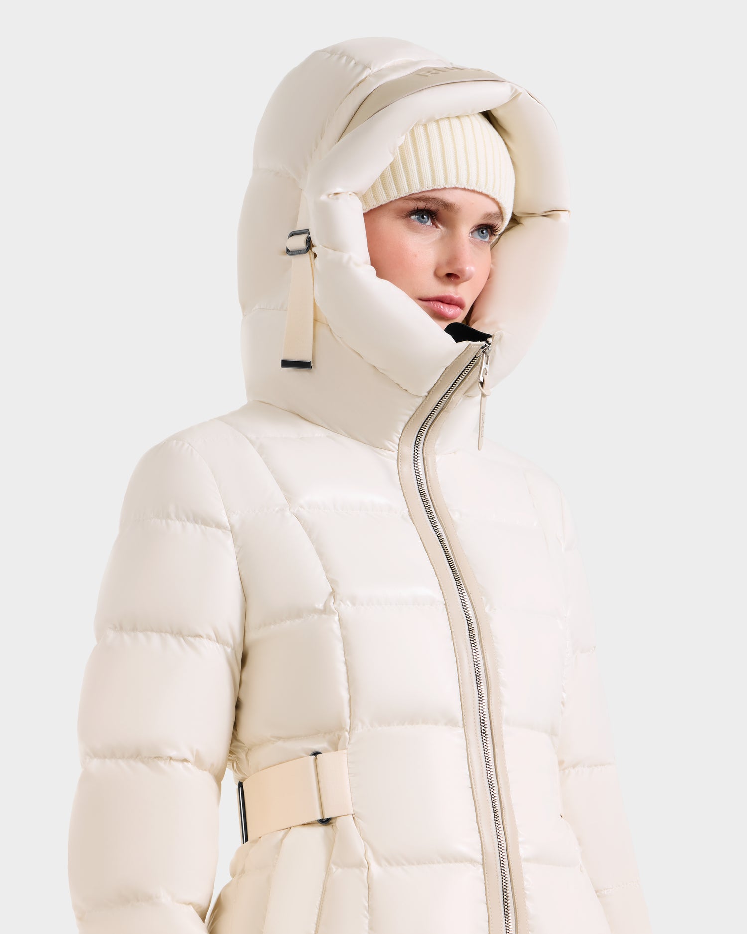 A woman wears the VANICE S - L124514 Vanilla knee-length down puffer by Rudsak with a large hood and beige knit beanie. The women's winter coat features a front zipper and side belt details as she looks to the side against a plain background.