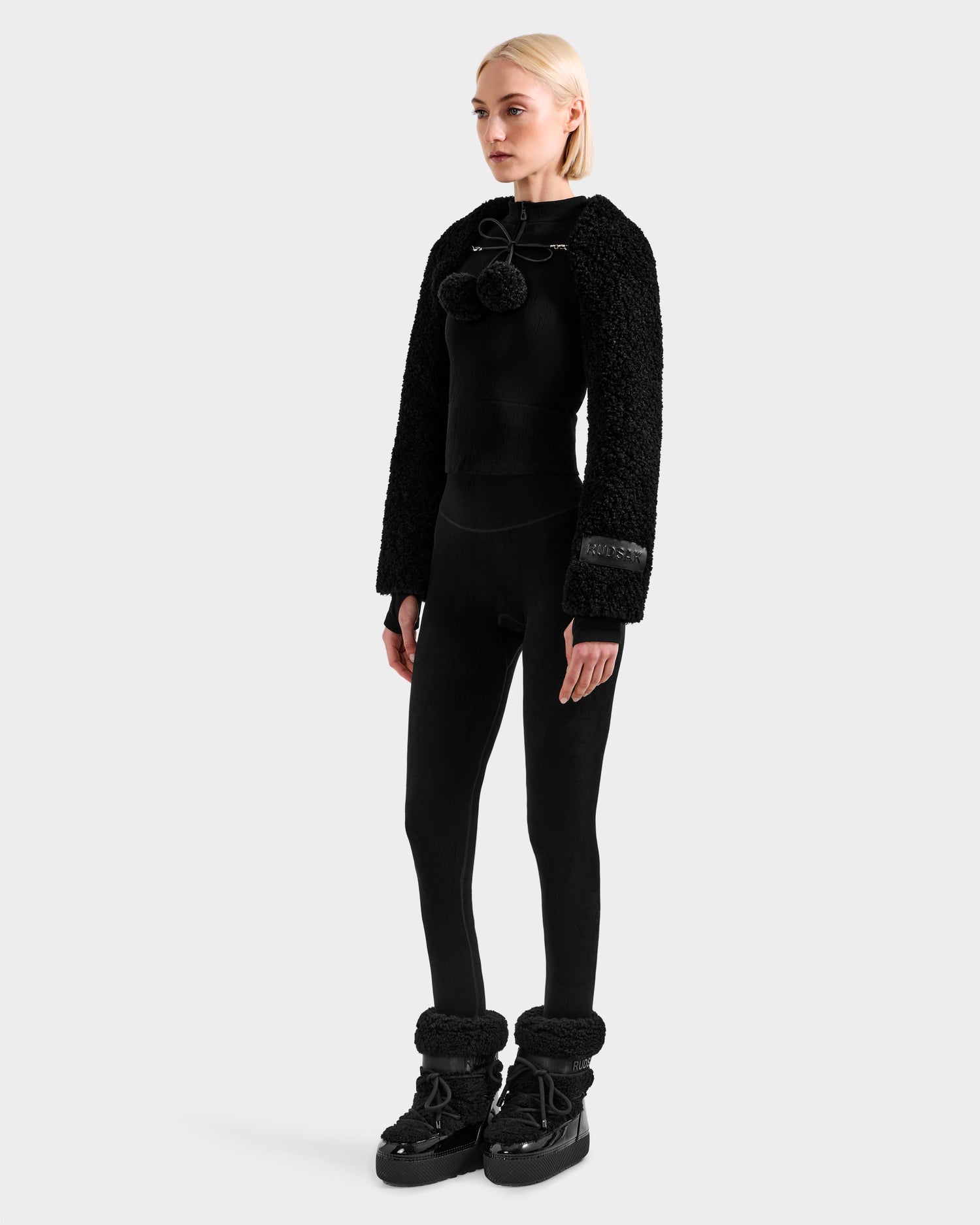 A woman with straight blonde hair and a neutral expression models Rudsak's VERITY - 8525666 Black: a women’s faux shearling bolero with pom-pom ties, paired with fitted leggings and black winter boots with furry cuffs.