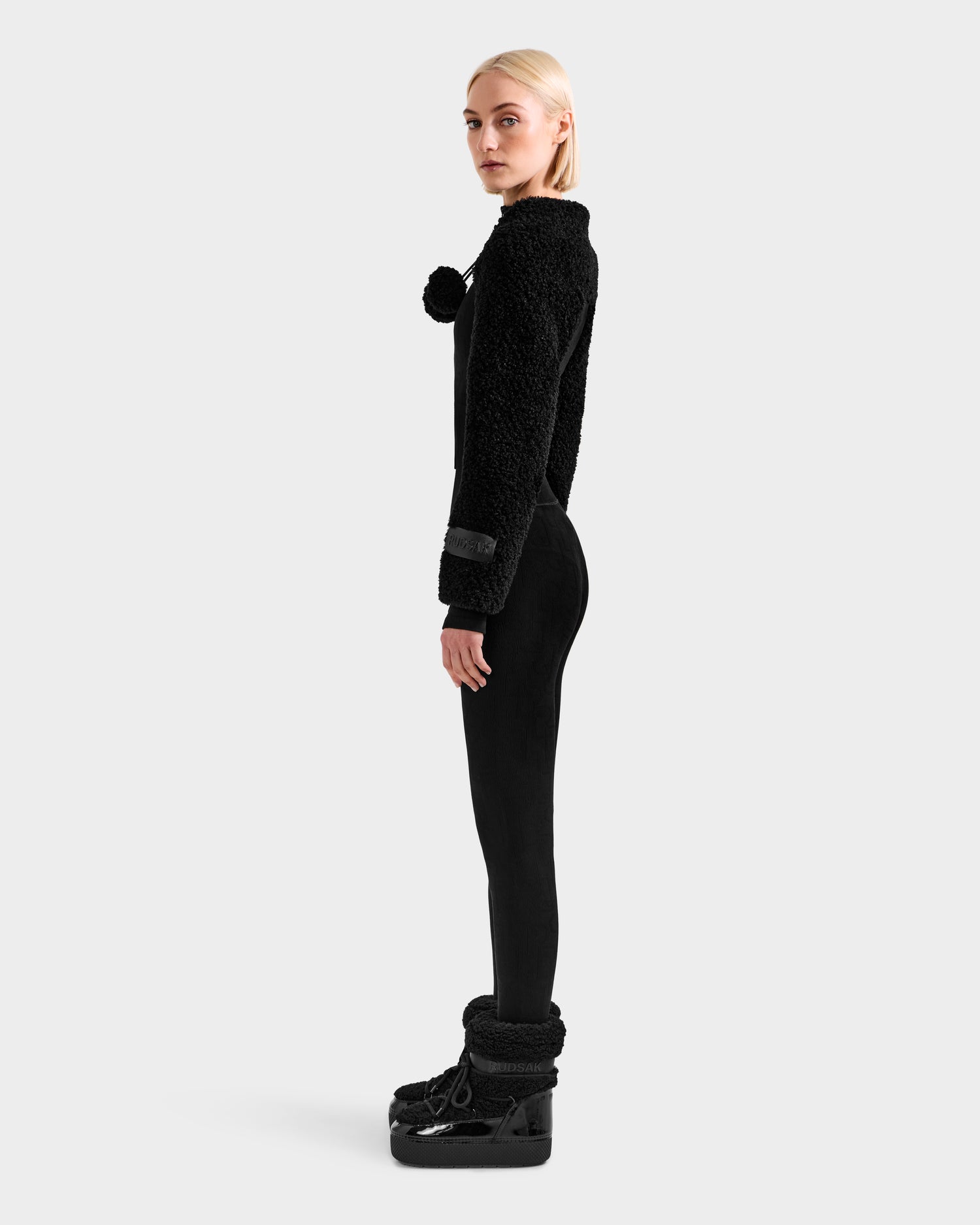 A woman with blonde hair stands in profile against a plain background, wearing the Rudsak VERITY - 8525666 Black faux shearling bolero over a black fuzzy sweater with black leggings and winter boots for a chic seasonal look.