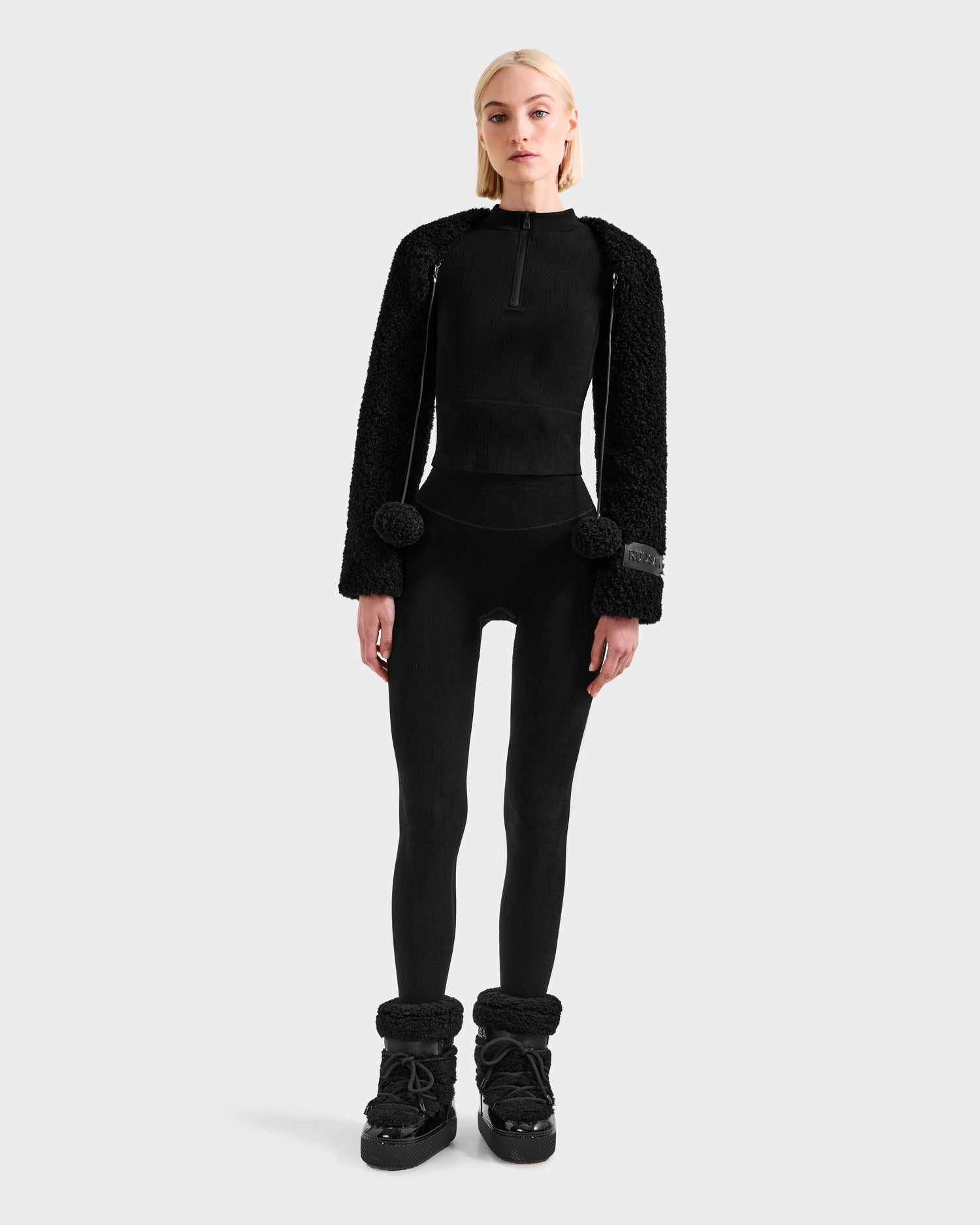A woman faces forward against a light gray background wearing Rudsak’s VERITY - 8525666 Black zip-up top, textured black statement outerwear with pom-poms, black leggings, and fur-trimmed black ankle boots.
