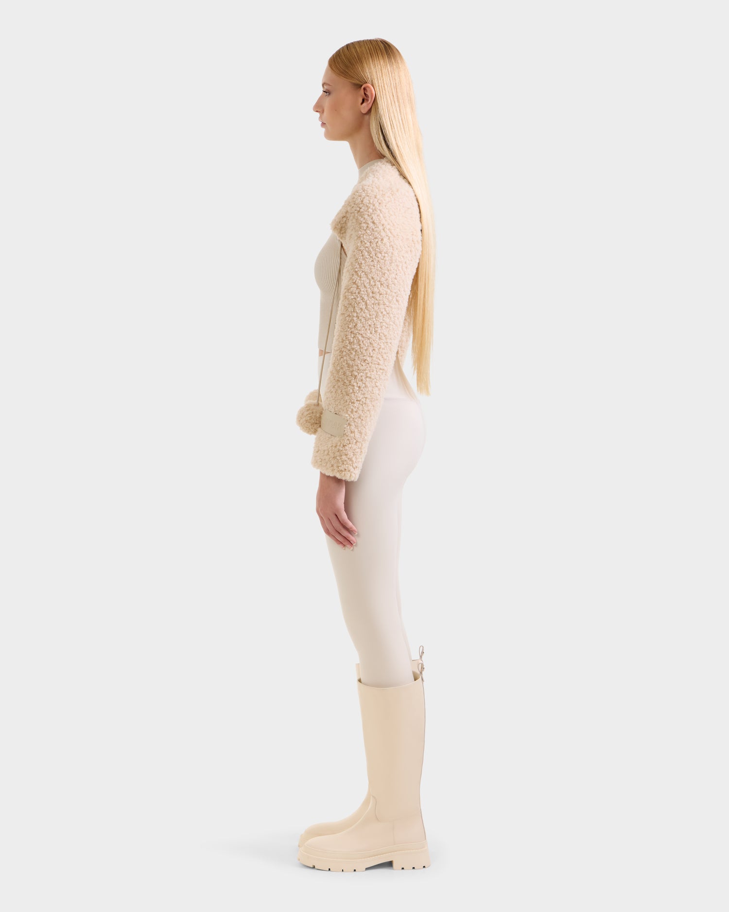 A woman stands in profile against a plain light background, wearing the Rudsak VERITY - 8525666 Vanilla women's faux shearling bolero cropped jacket, paired with fitted light pants and matching knee-high boots for an effortlessly chic winter look.