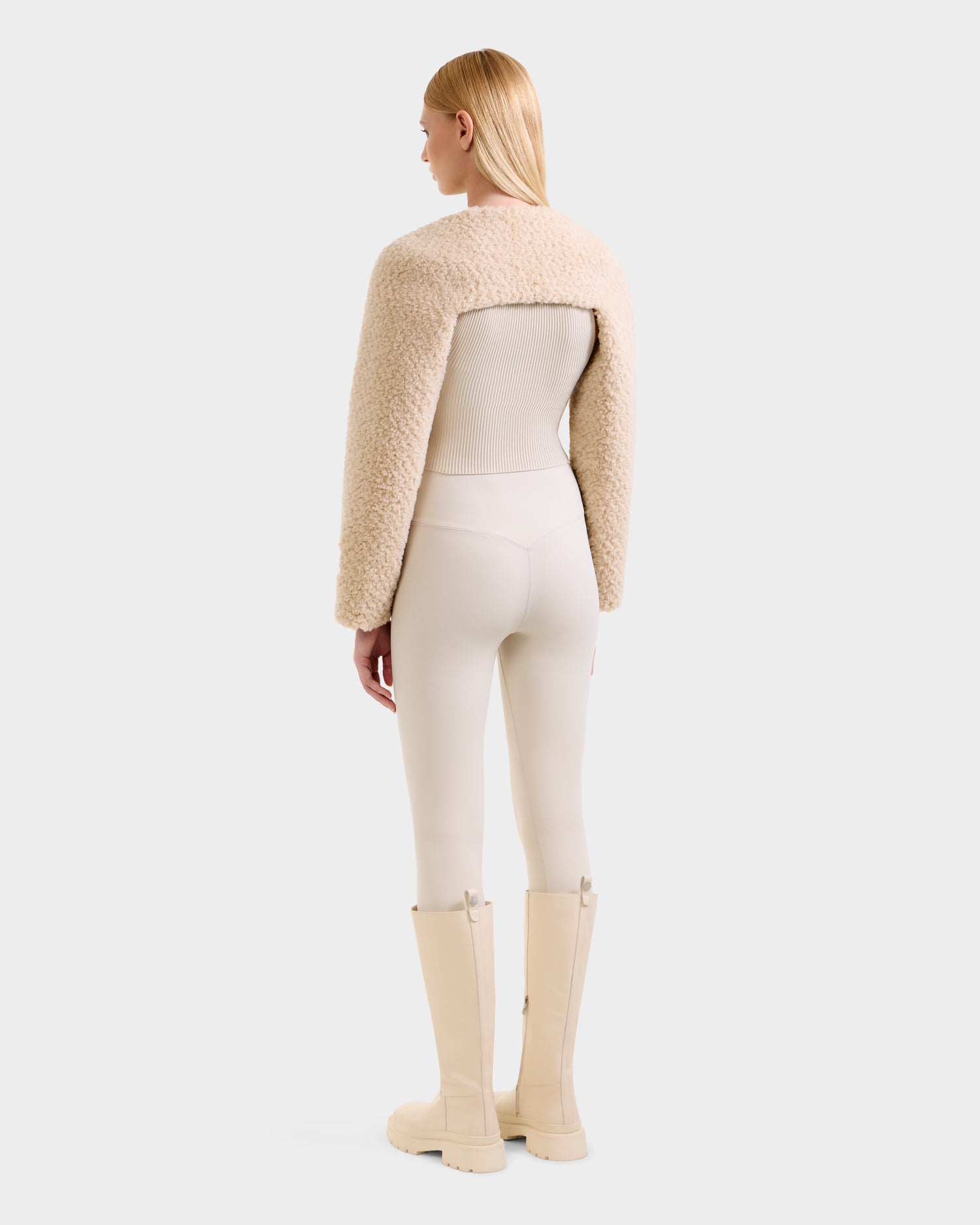 A woman, seen from behind, wears Rudsak’s VERITY - 8525666 Vanilla faux shearling bolero with a cream ribbed top, matching leggings, and beige knee-high boots—a chic après-ski look against a plain light gray background.