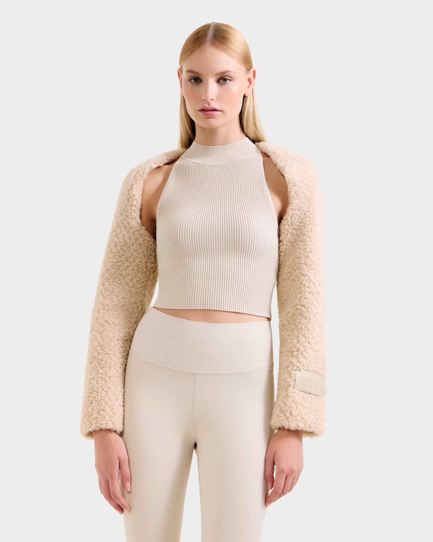 A woman with straight blonde hair wears the VERITY - 8525666 Vanilla faux shearling bolero by Rudsak, styled with a sleeveless beige crop top and high-waisted leggings, standing against a plain light gray background.
