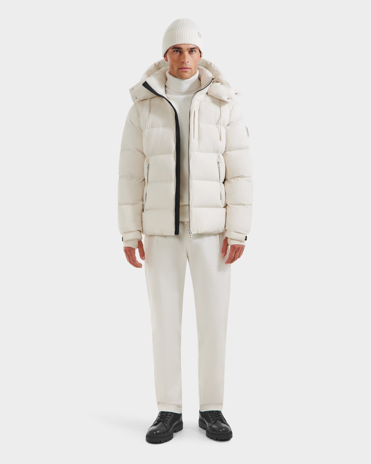 A person wearing the VINCE M - 6124561 Vanilla mens down puffer jacket by Rudsak, with RDS-certified down, a white turtleneck, white pants, white beanie, and black shoes stands against a plain white background.