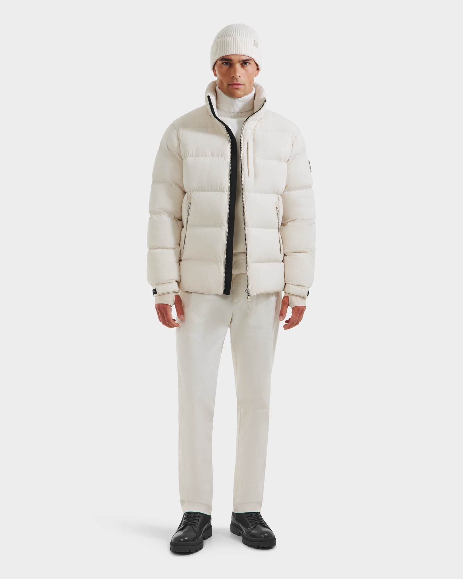 A man faces forward wearing the Rudsak VINCE M - 6124561 Vanilla quilted down puffer jacket, styled with a white turtleneck, white pants, white knit beanie, and black shoes against a plain white background.