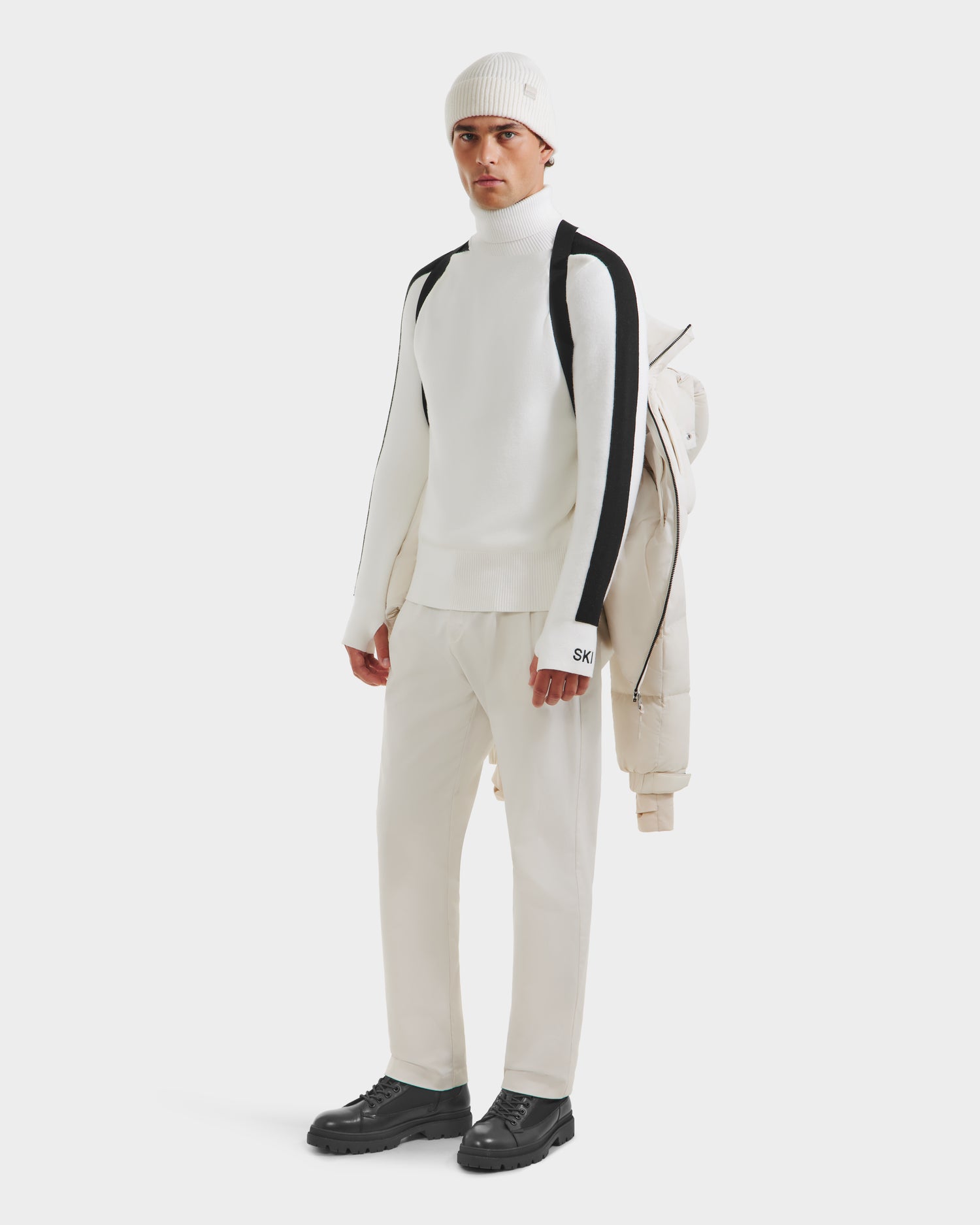 A man stands against a plain background wearing Rudsak’s VINCE M - 6124561 Vanilla: a white turtleneck sweater with black sleeve stripes, cream pants, black shoes, a white beanie, and holding a weather-resistant shell jacket over his shoulder.