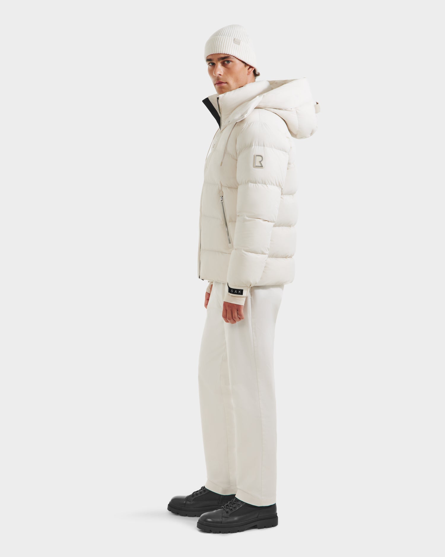 A person in the Rudsak VINCE M - 6124561 Vanilla quilted down puffer jacket with hood, a white beanie, white pants, and black shoes stands sideways on a plain white background looking forward.