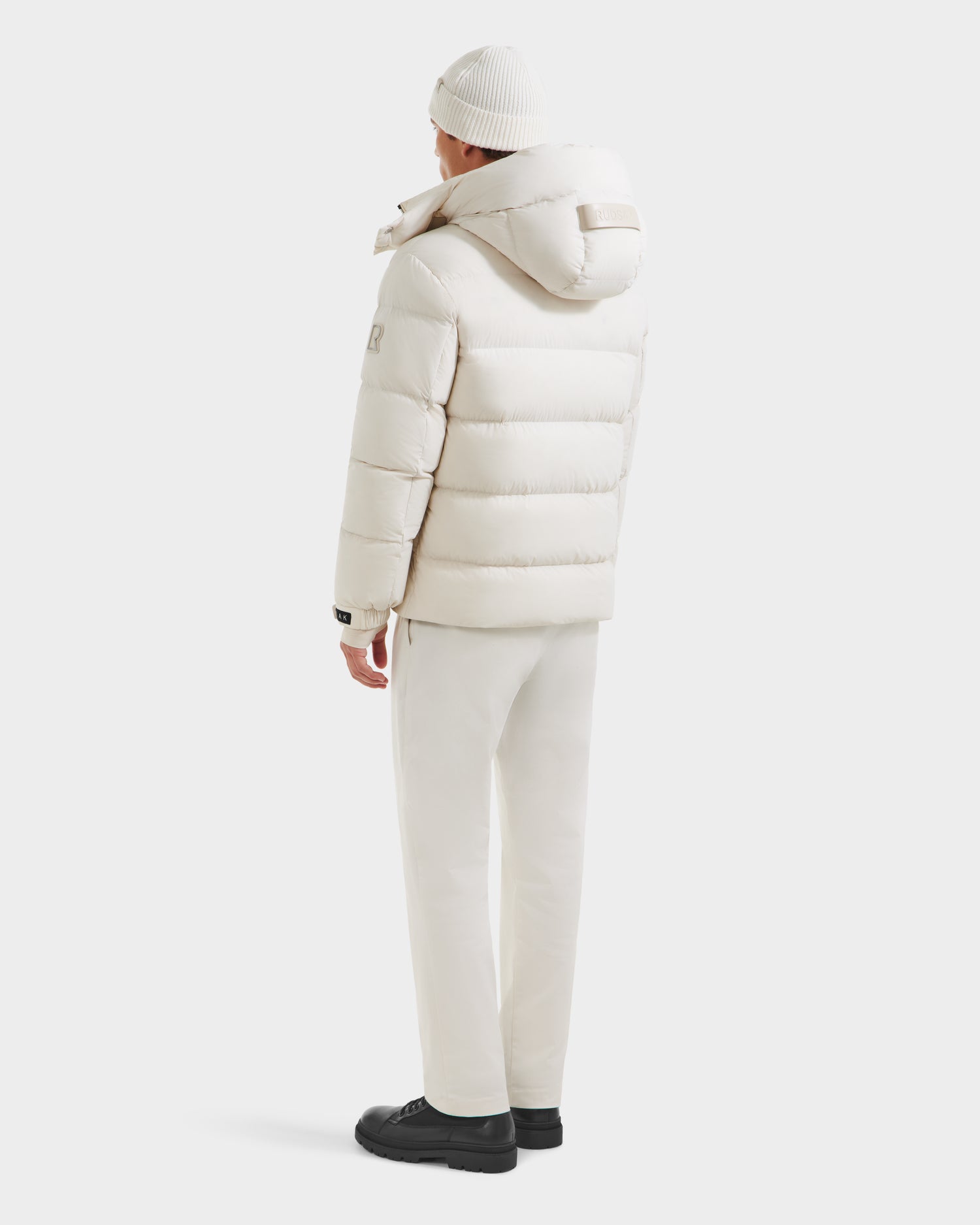 Dressed in the Rudsak VINCE M - 6124561 Vanilla men’s quilted down puffer jacket, matching pants, beanie, and black shoes, a person stands facing away against a white background, hands relaxed at their sides.