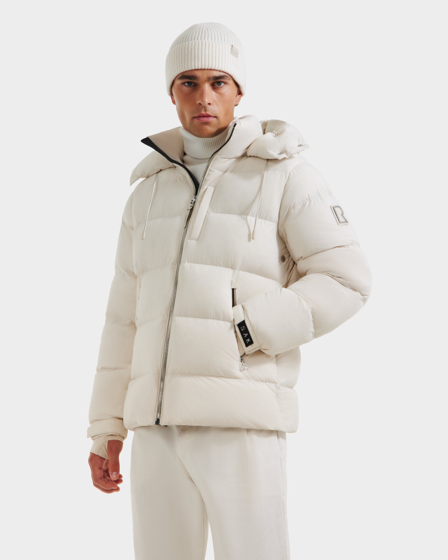 A man in an all-white winter outfit, featuring the Rudsak VINCE M - 6124561 Vanilla men’s down puffer jacket, a beanie, turtleneck sweater, and pants, stands against a plain white background.